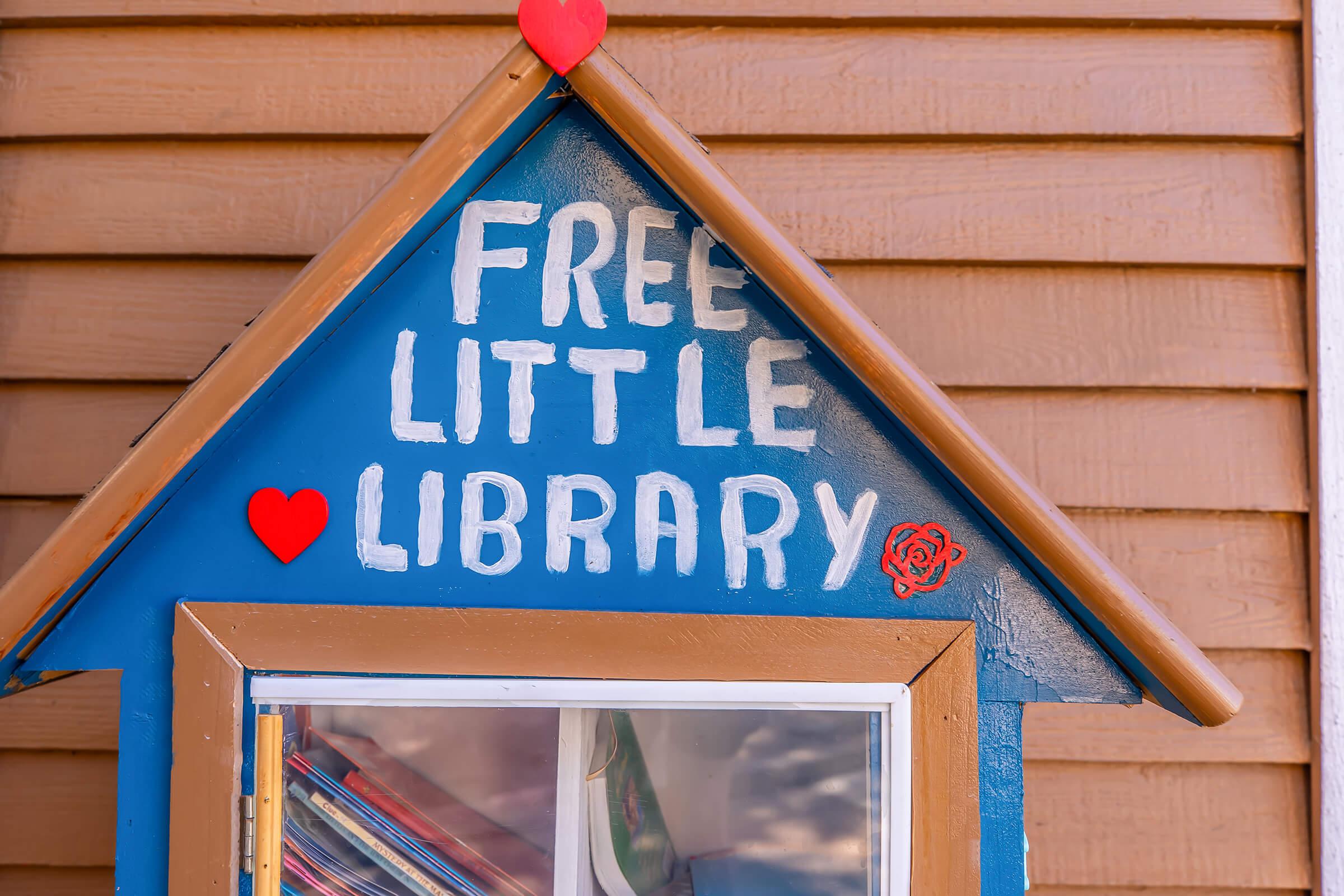 A small, colorful "Free Little Library" box with a peaked roof. The sign features white text on a blue background and is adorned with red hearts. The box is inviting and encourages community book sharing. Nearby wooden siding is also visible in the background.