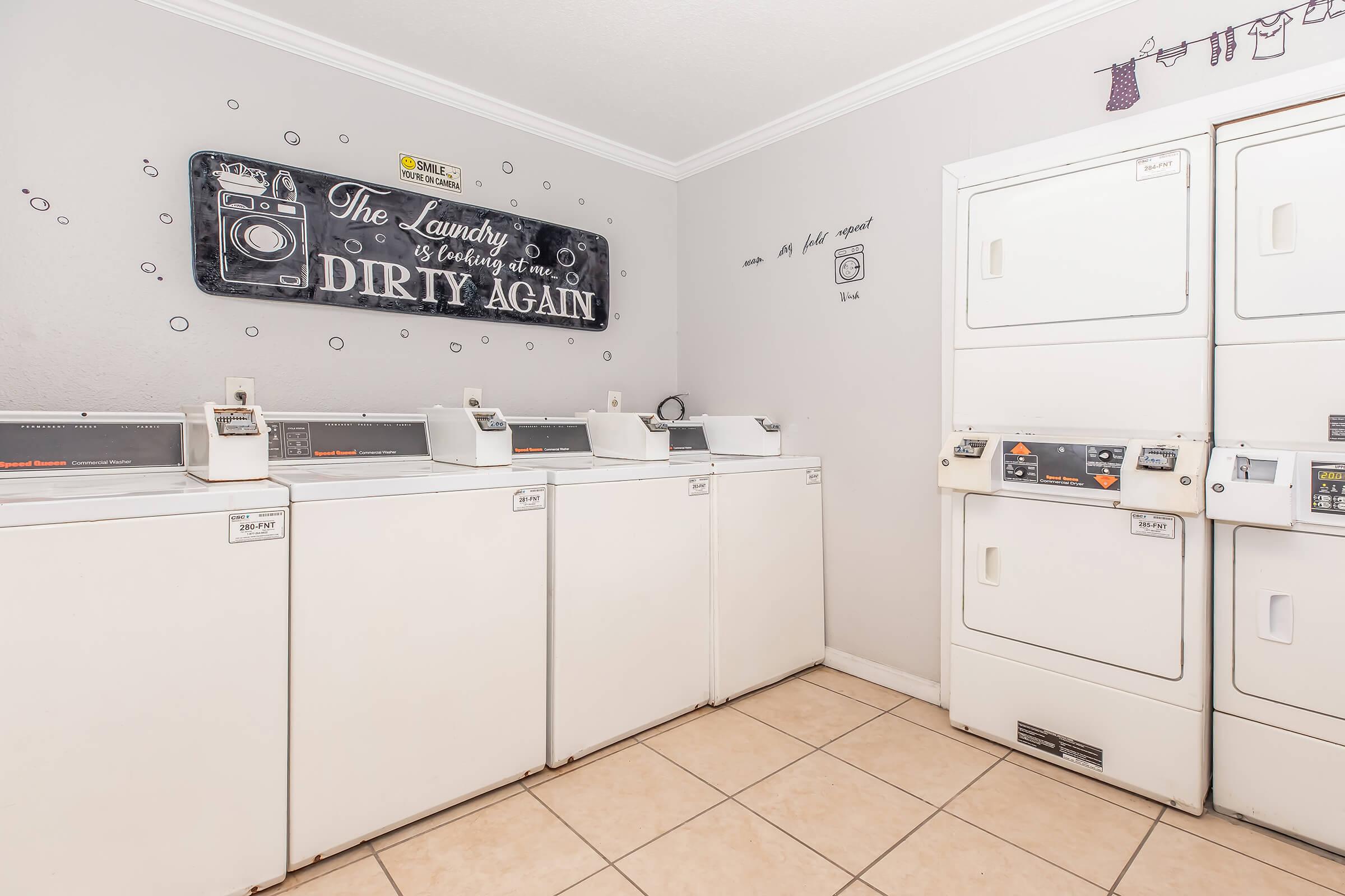 A bright laundry room featuring several white washing machines and dryers. The walls are decorated with playful wall art, including a sign that reads "The laundry is calling me, DIRTY AGAIN." The floor is tiled, and the overall ambiance is clean and inviting.