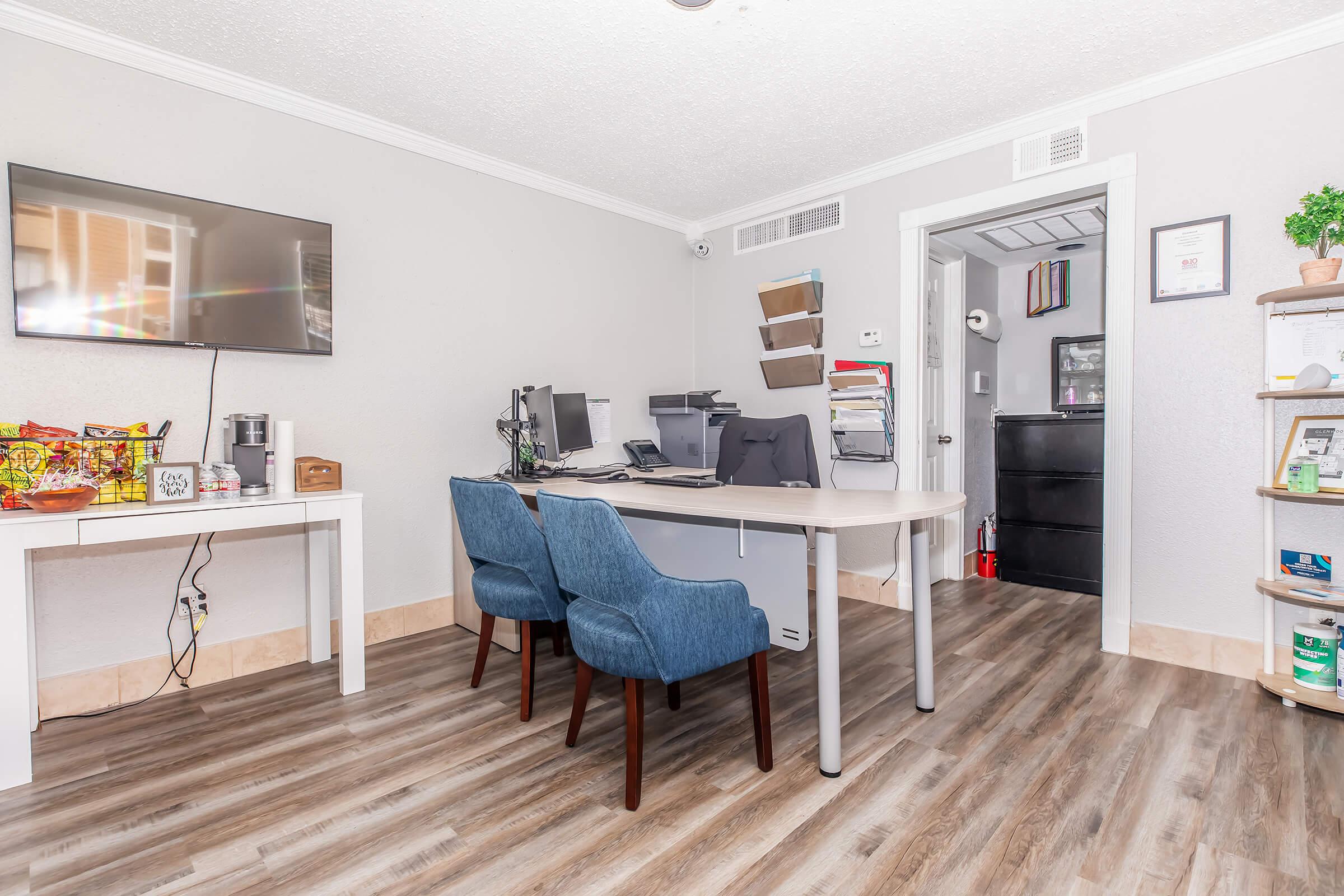 A modern office interior featuring a wall-mounted TV, two blue chairs at a white desk, and two computer workstations. A storage cabinet is visible in the background, along with shelves displaying various items and snacks. The flooring is wood-like, creating a warm, inviting atmosphere.