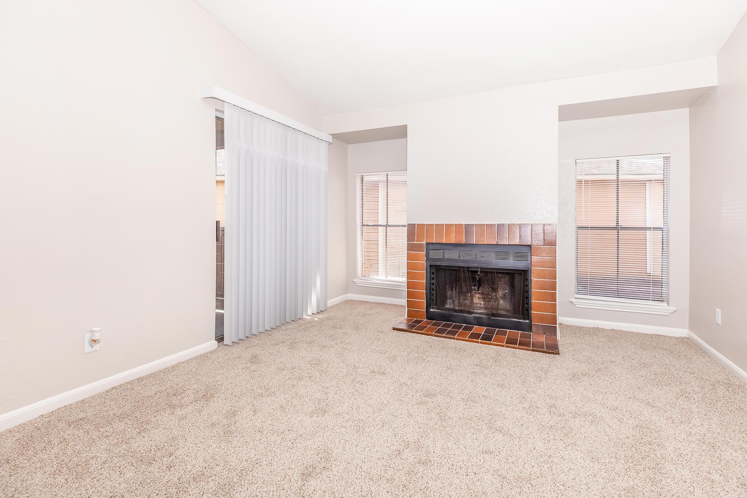 A spacious living room featuring a tan carpet, a fireplace with a brown tiled surround, and large windows with blinds. One wall includes sheer white curtains, and the overall color scheme is neutral, creating a bright and inviting atmosphere.