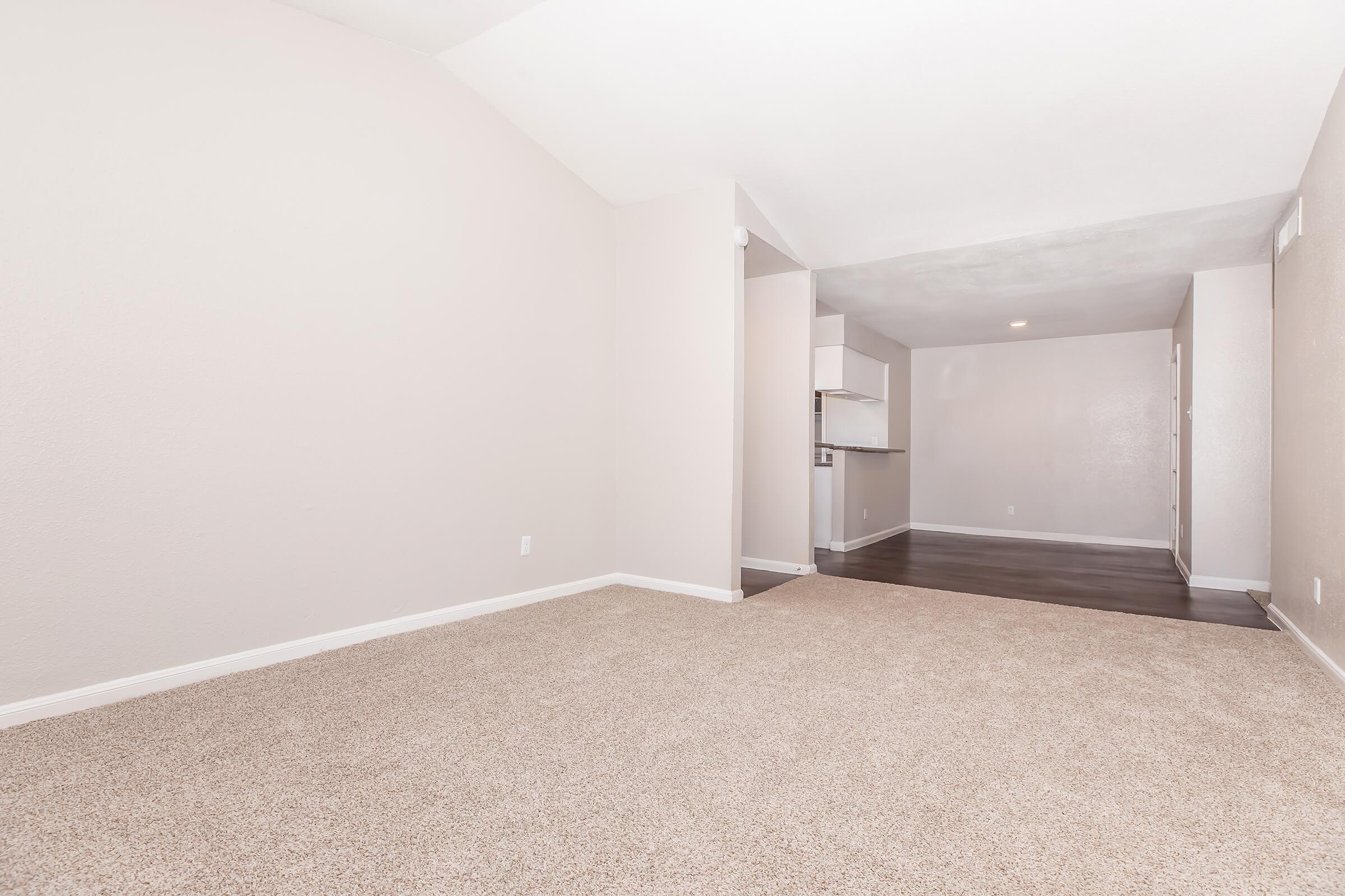 Empty living space with light beige carpet and soft pink walls. The room features a vaulted ceiling and leads to a kitchen area in the background, which has dark wood flooring. There are no furnishings in the room, creating an open and spacious feel.
