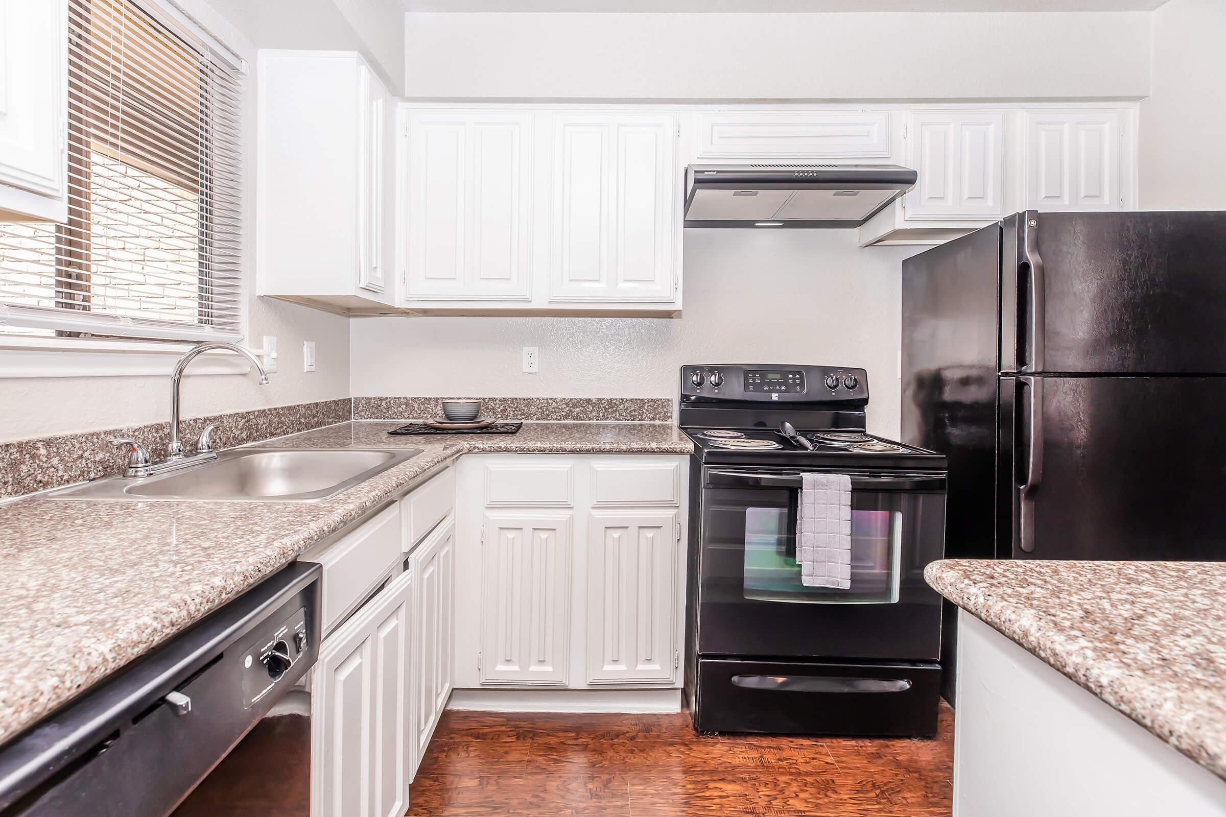 A modern kitchen featuring white cabinets, a black refrigerator, and a black stove with an oven. The countertops are a speckled granite, and there is a sink on the left side. The room has a bright atmosphere with a window allowing natural light.