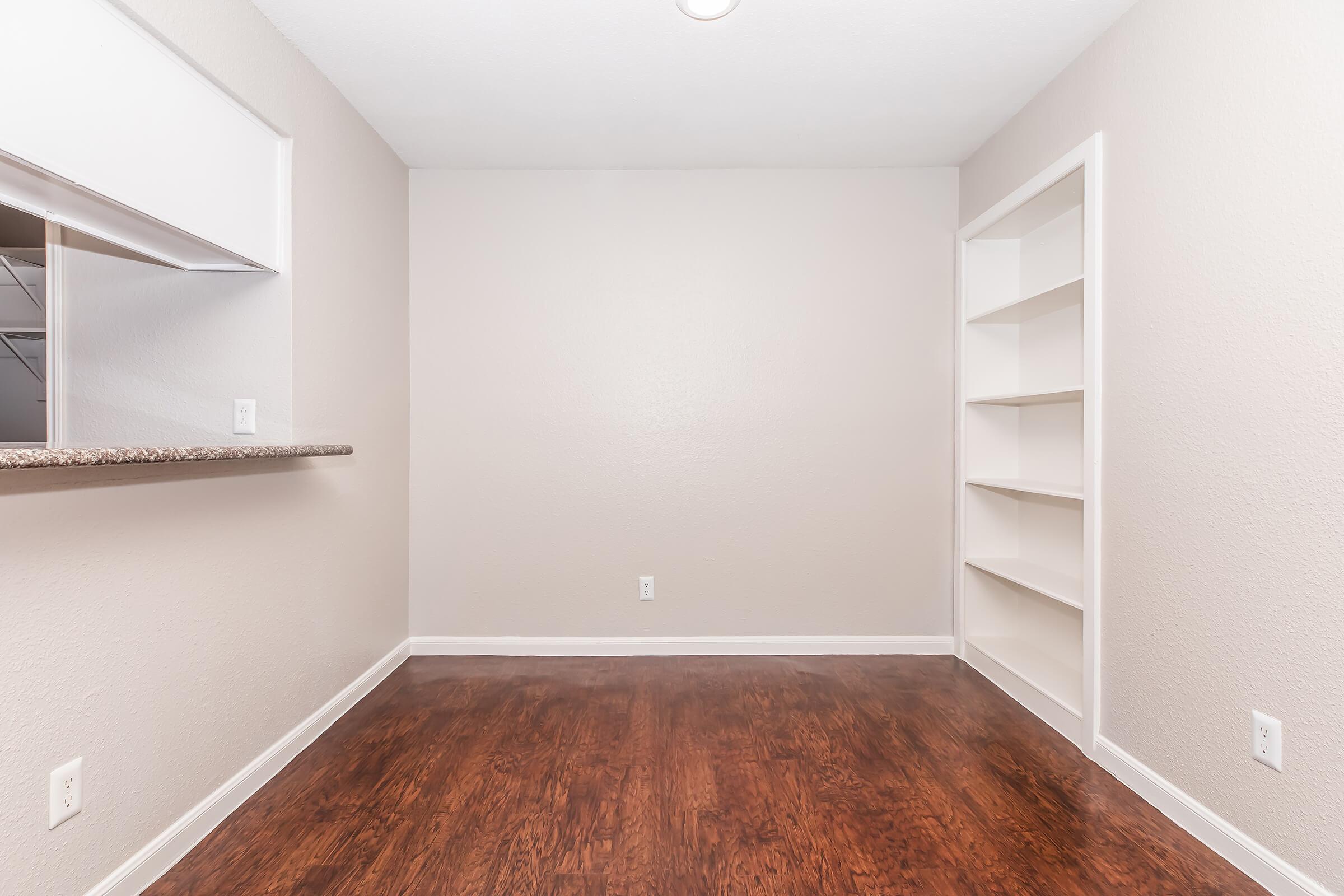 A small, empty room with light beige walls and dark wooden flooring. There is a window on one side with a counter attached, and a built-in shelf along the adjacent wall. The space feels open and minimalistic, suitable for various uses such as an office or reading nook.