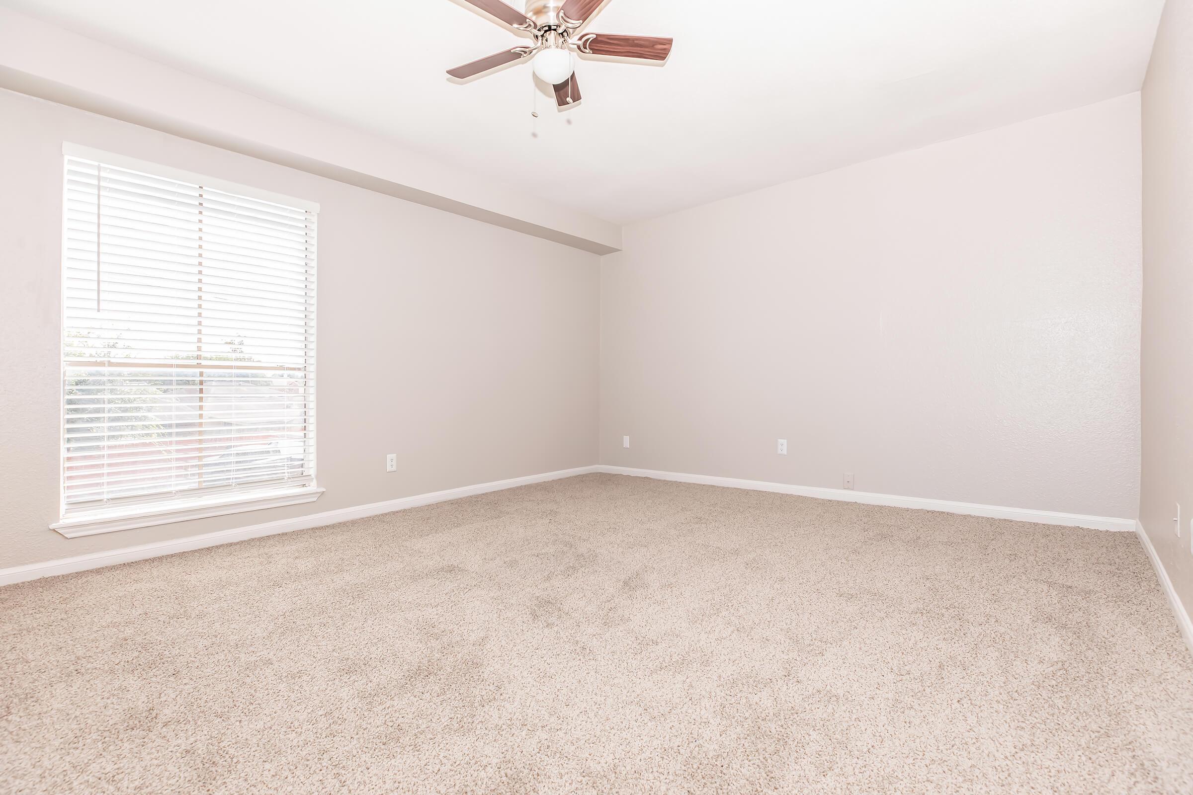 A spacious, empty room with light beige walls and a ceiling fan. The floor is covered in plush, light-colored carpet. There is a large window with white blinds allowing natural light to fill the space. The room appears clean and well-maintained, providing a neutral backdrop for personal decoration.