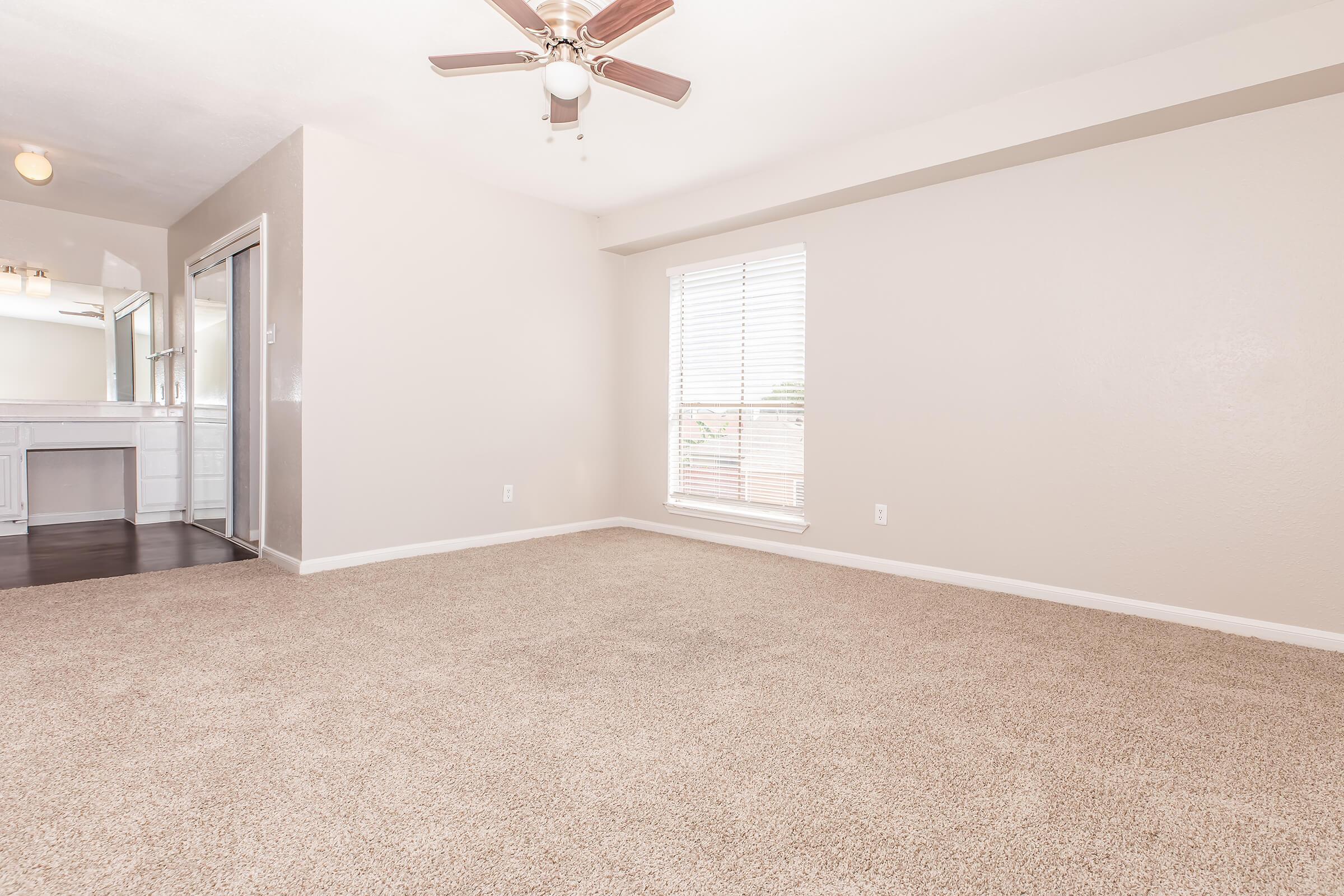 Spacious, empty living room with light beige carpet, a ceiling fan, and a window with white blinds. A mirror and sink area are visible in the background, adding to the bright and airy feel of the space. The walls are painted in a soft neutral tone, enhancing the openness of the room.
