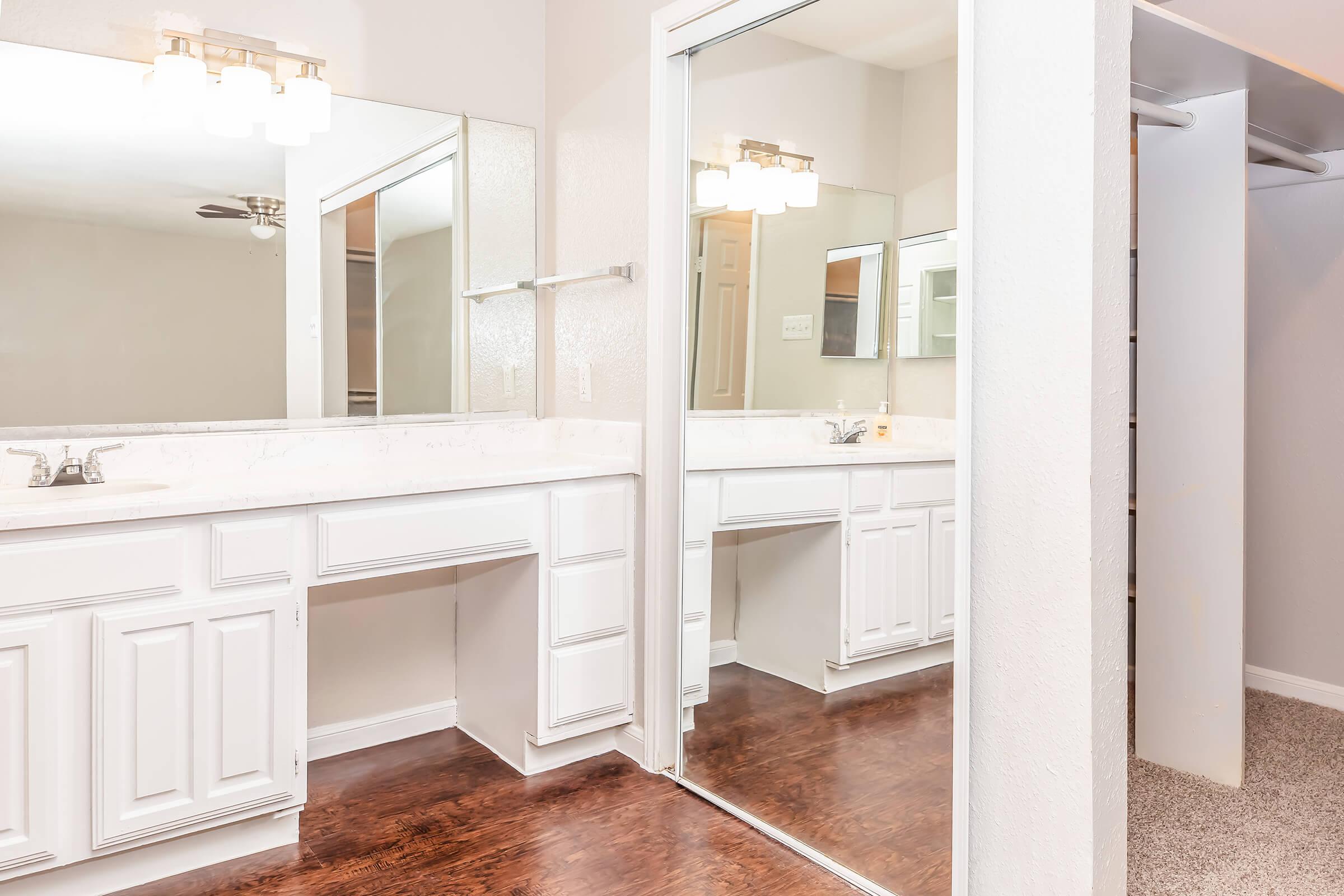 A modern bathroom featuring double sinks with a white vanity, large mirrors, and stylish lighting. A glass shower enclosure is visible, along with a closet area. The floor has a warm wood finish, contrasting with the light-colored walls and fixtures. Overall, the space is bright and inviting.