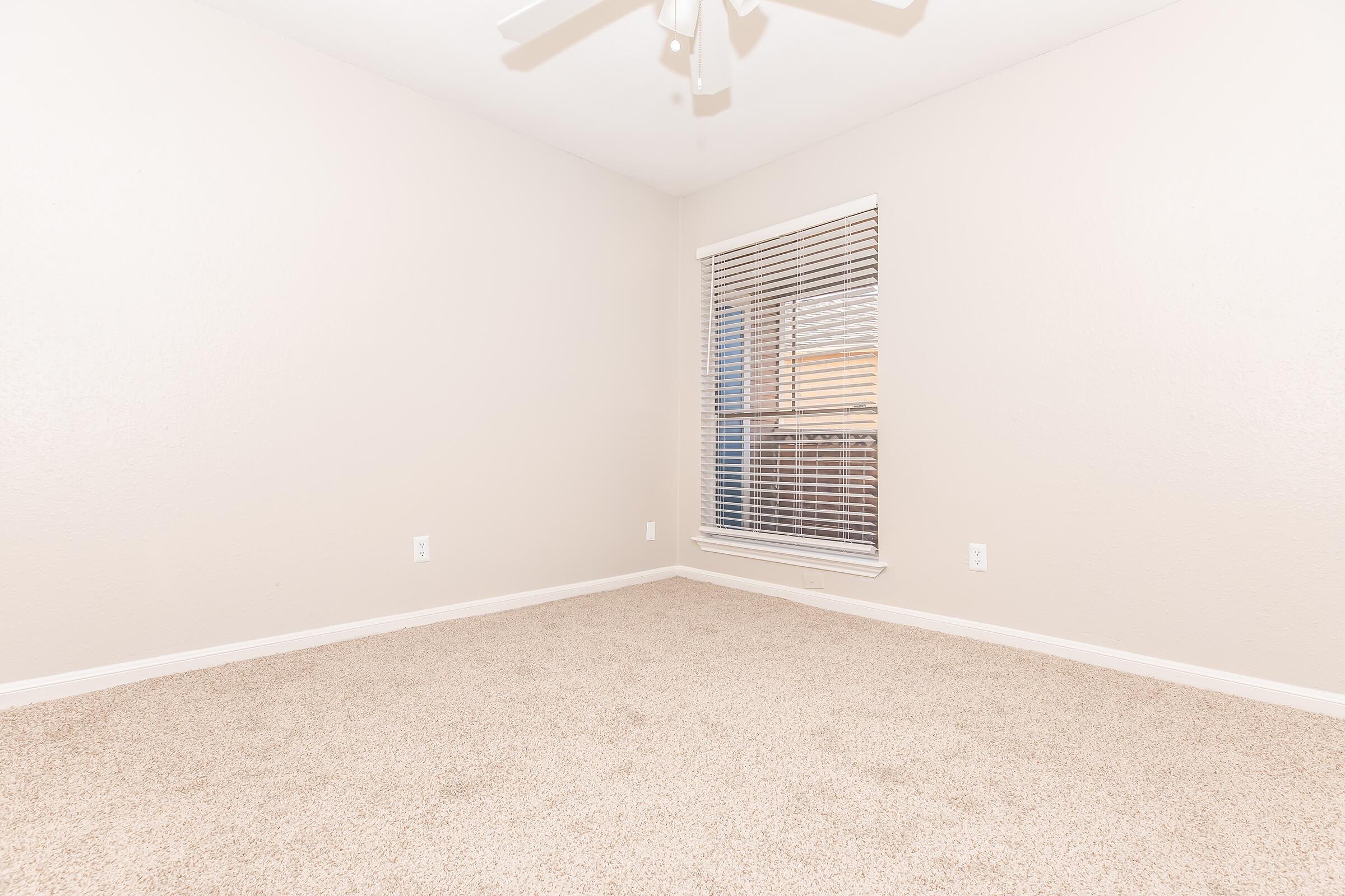 An empty room with light beige walls and carpet, featuring a window with white blinds. The room has a ceiling fan and is well-lit, creating a spacious and clean atmosphere.