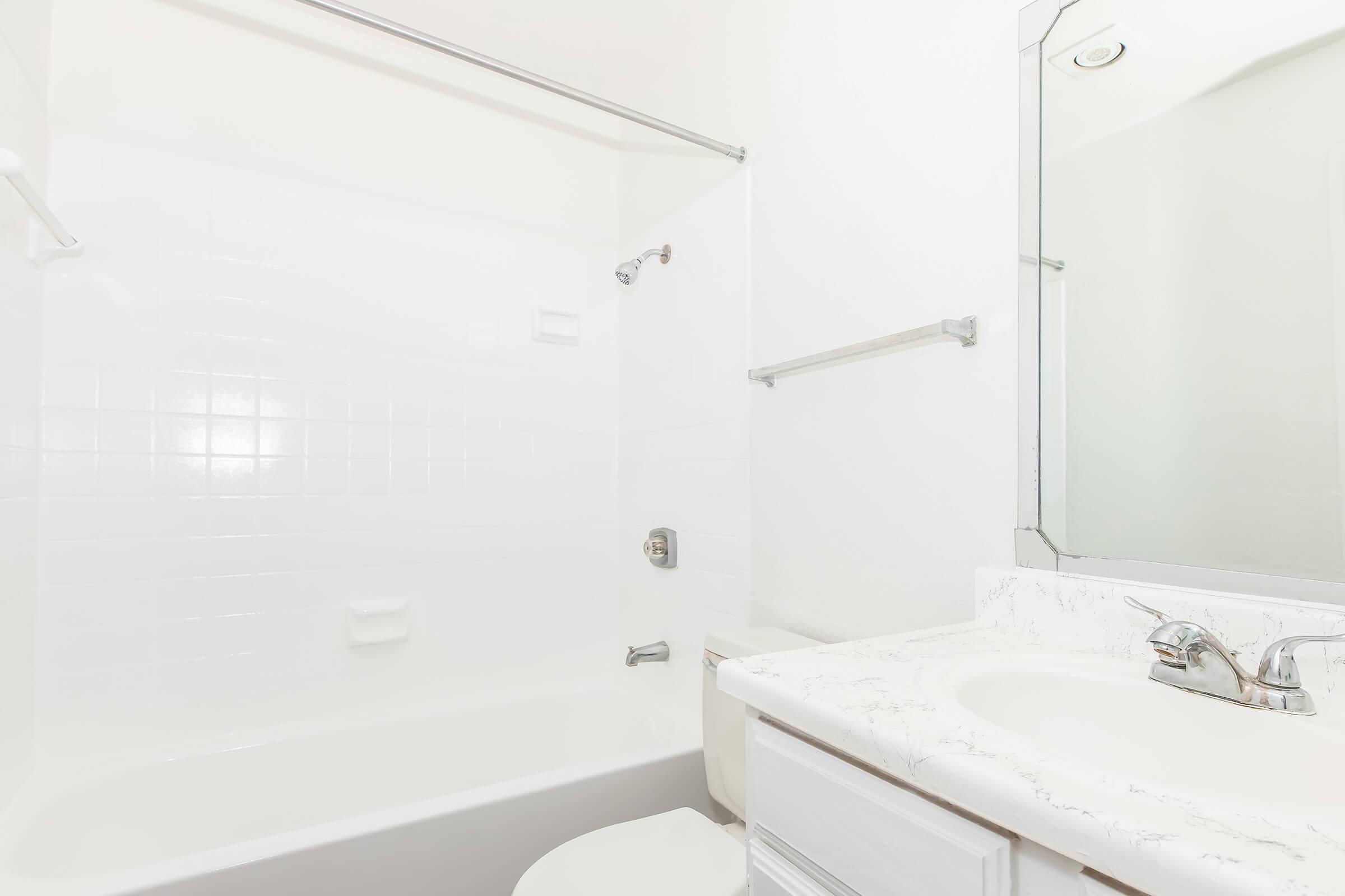 A clean, simple bathroom featuring a white tiled bathtub and shower area, a countertop with a mirror above, and a sink. The walls are painted white, providing a bright and airy feel. A towel bar can also be seen on the wall.