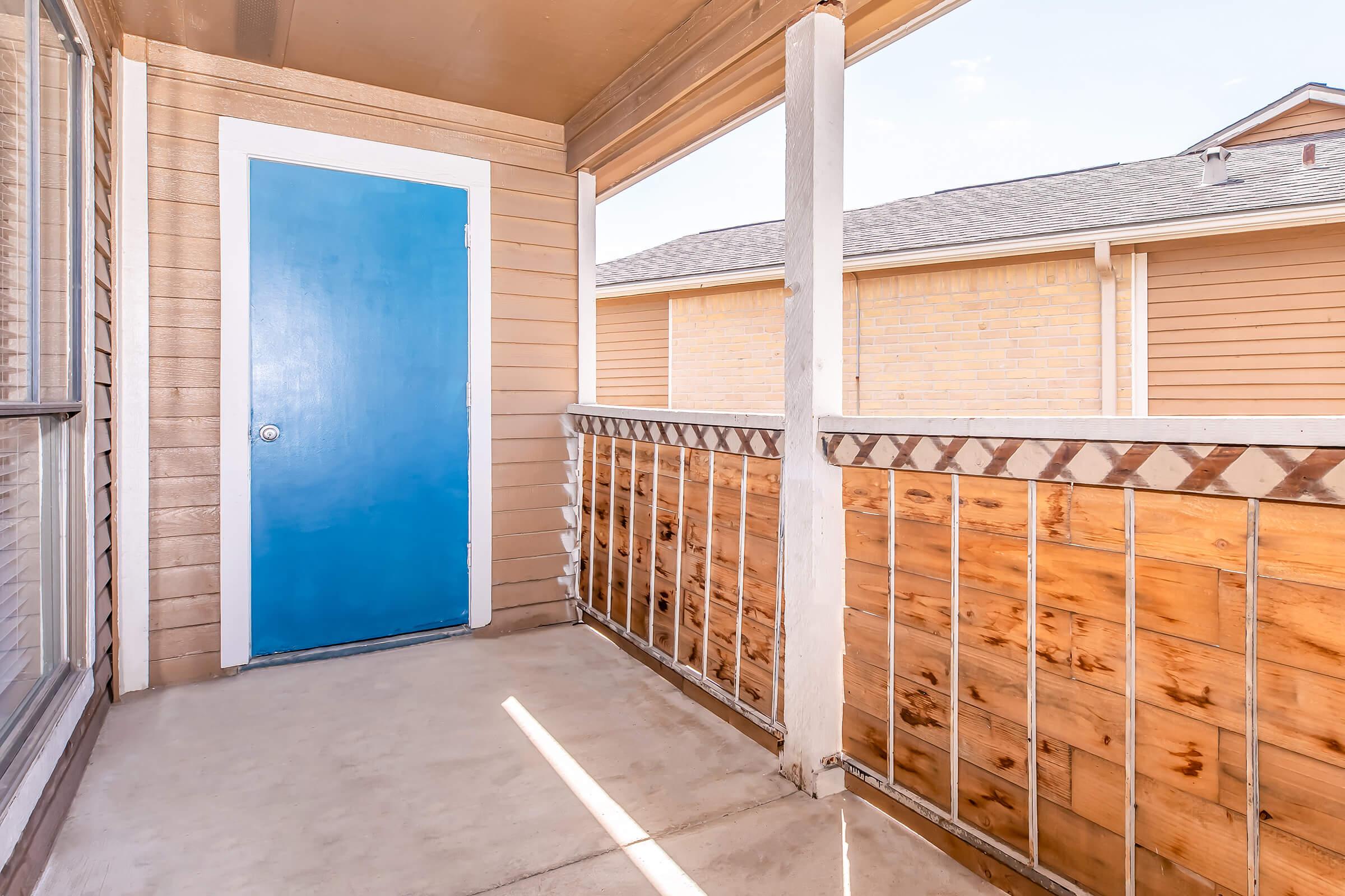 A small balcony area with a bright blue door, wooden railings, and beige siding. The floor is concrete, and there is natural light coming in from the side, illuminating the space. The view includes a neighboring building with a simple brick exterior.