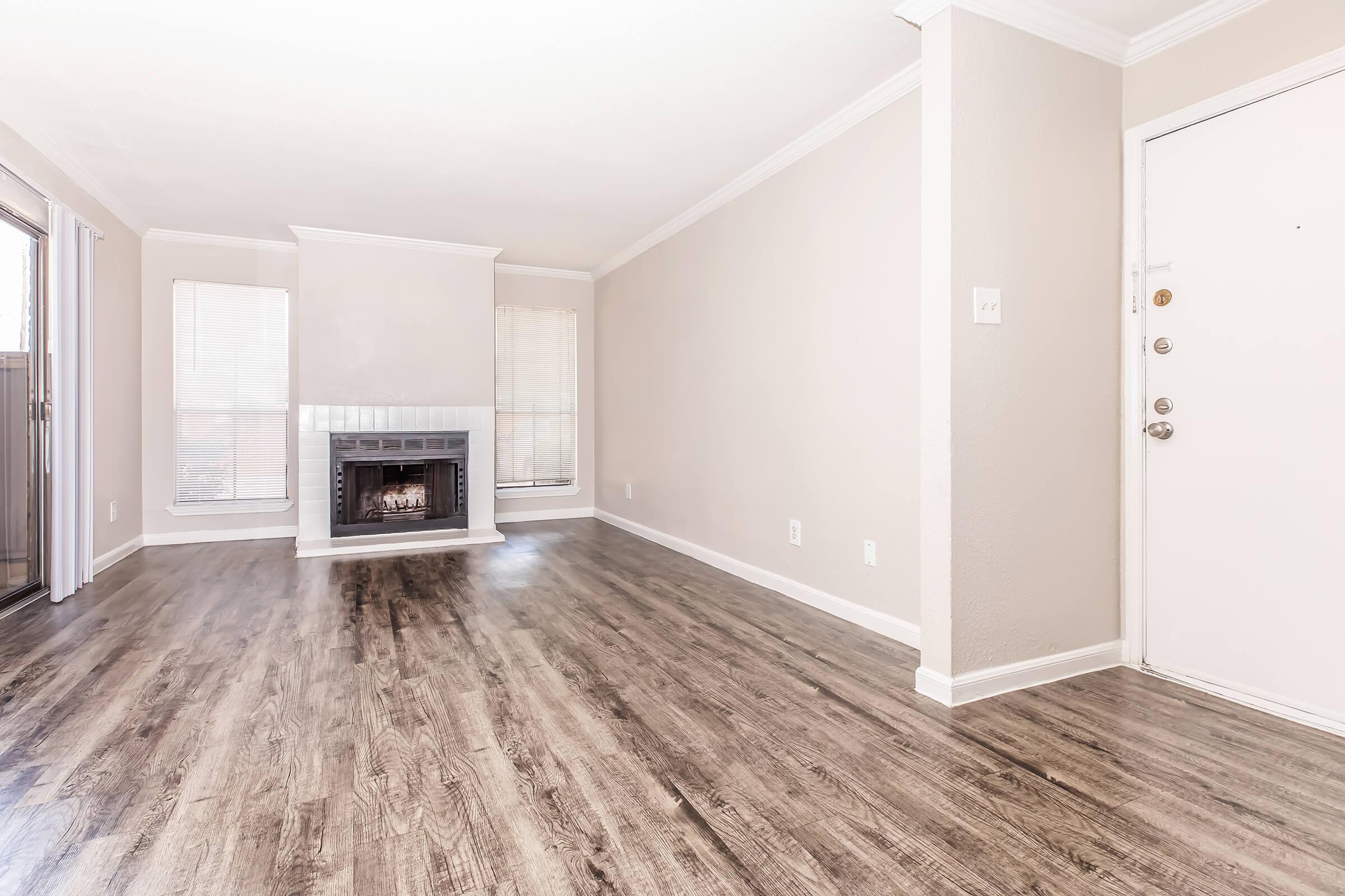 A well-lit living room featuring a light-colored wall, large windows with blinds, and a modern fireplace. The floor is covered in sleek, dark wood-like laminate. A white front door is visible on the right side, and the space is free of furniture, creating an open and inviting atmosphere.