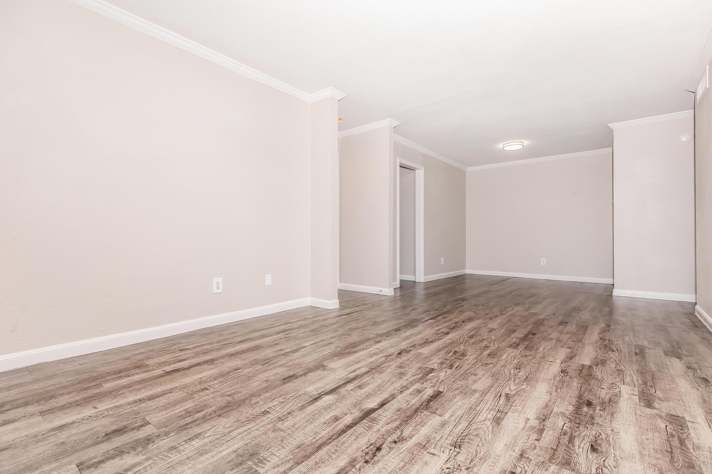 An empty, well-lit room with light-colored walls and a wooden floor. The space features a simple ceiling light, baseboards, and an open doorway leading to another area. The overall atmosphere is clean and minimalistic, ideal for various uses.