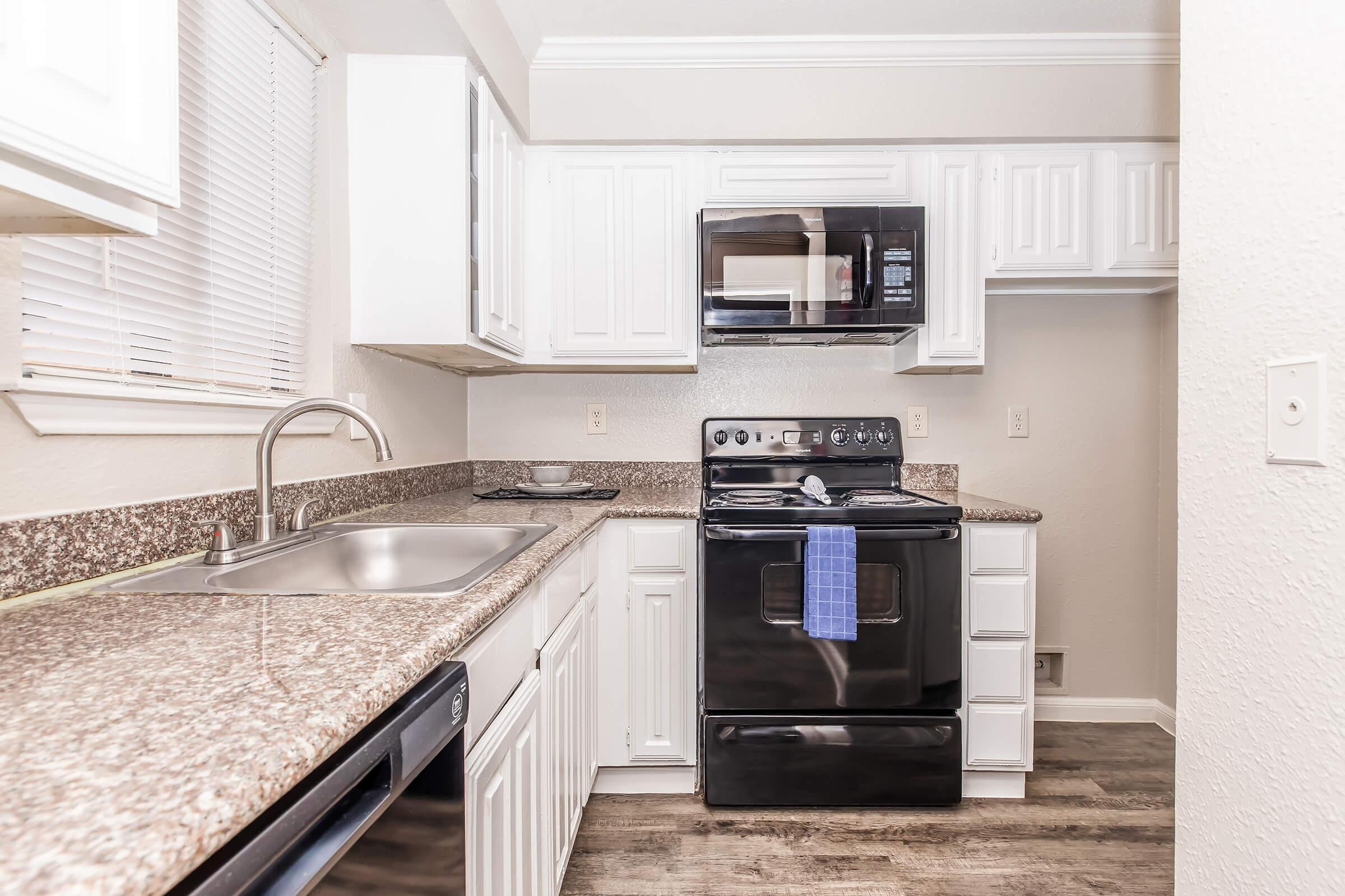 Modern kitchen featuring white cabinetry, a black stove and microwave, a stainless steel sink, and a stone countertop. Natural light streams in through a nearby window, enhancing the spacious feel of the room. Clean and contemporary design with a focus on functionality.