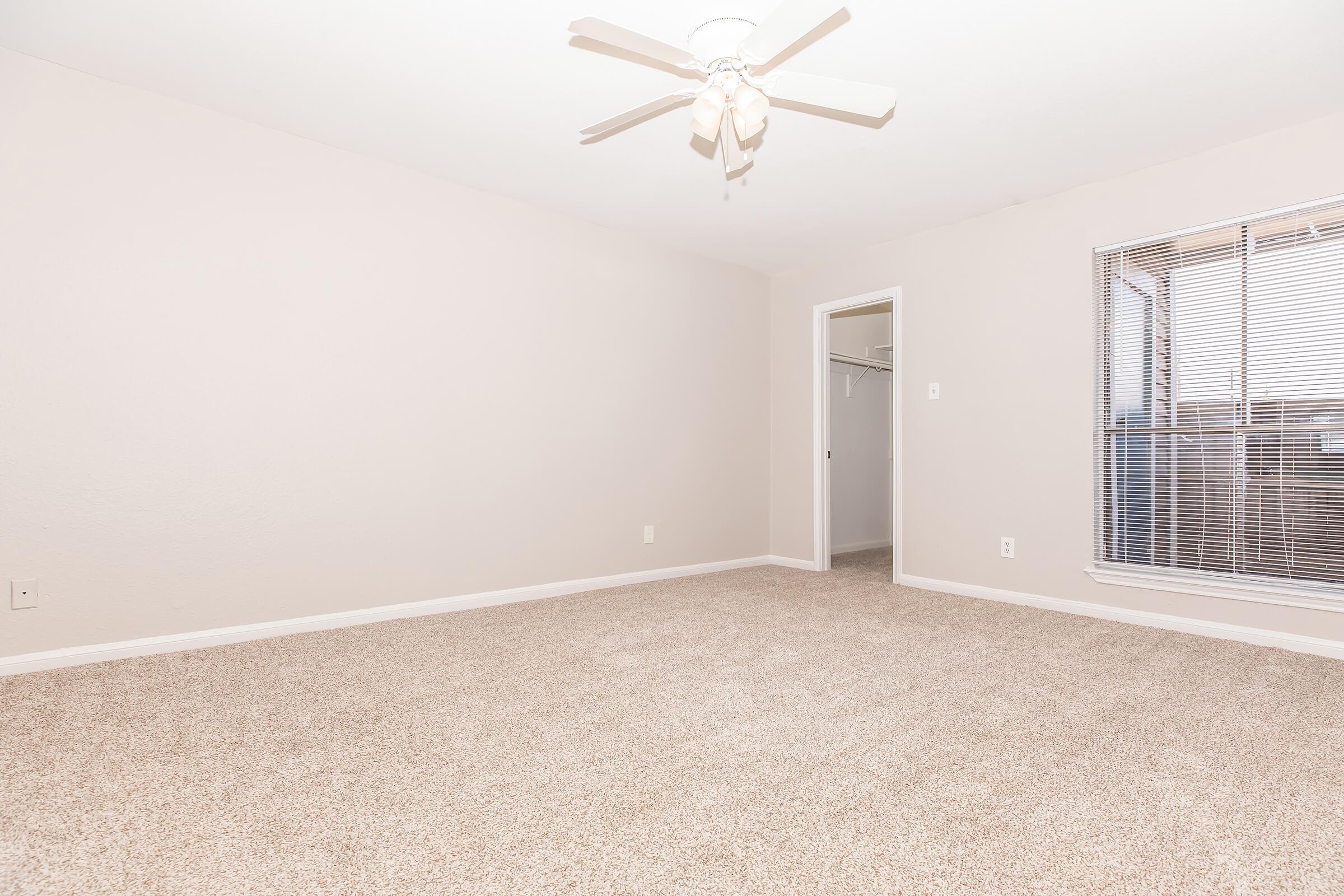 A spacious, neutral-colored room with light beige walls and a ceiling fan. The carpeted floor is plush and light. A window with vertical blinds lets in natural light, and there is an open door leading to a closet space. The overall atmosphere is bright and inviting, perfect for various uses.