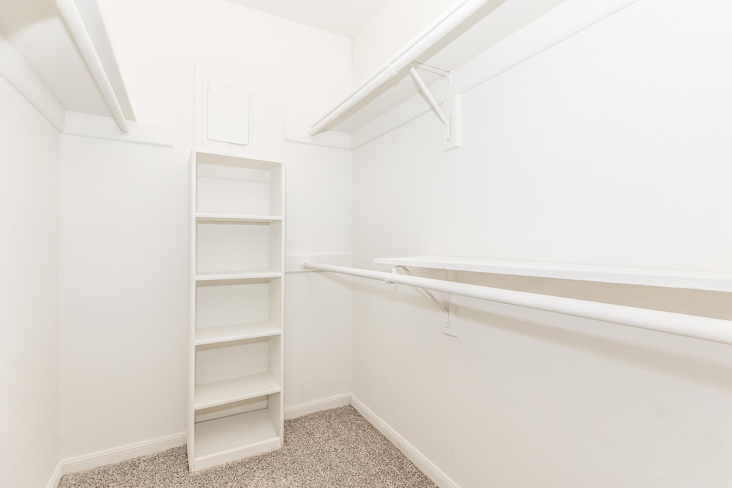 A clean, empty walk-in closet featuring white walls and shelves. There is a tall white shelving unit on one side and two hanging rods along the back wall, providing ample storage space. The floor is covered with carpet, adding a cozy feel to the minimalist design.