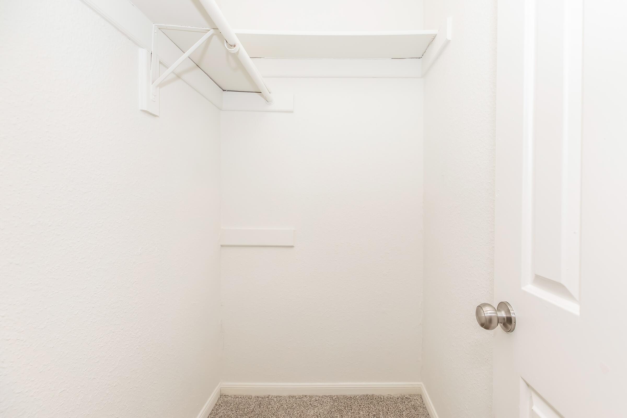 A small, empty closet with white walls and a light-colored carpet. The closet features a single shelf along the top and a door with a silver handle. The overall look is clean and minimalistic, with no items or decorations visible inside.