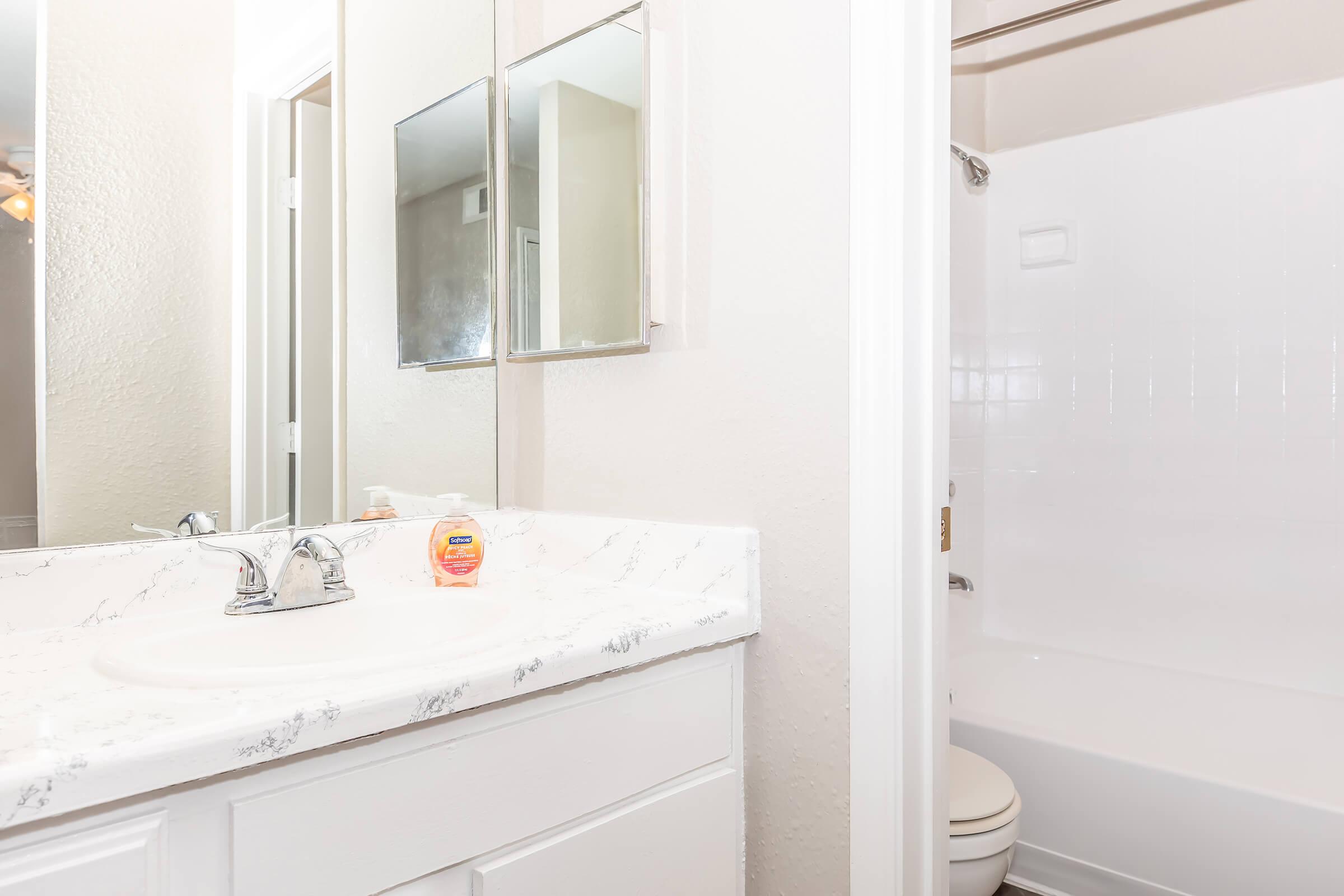 A clean bathroom featuring a white countertop with a sink and chrome faucets. Two mirrors are mounted above the sink. A shower/tub combination is visible to the right, with white walls and a light, neutral color scheme throughout the space. Soft lighting enhances the airy feel.