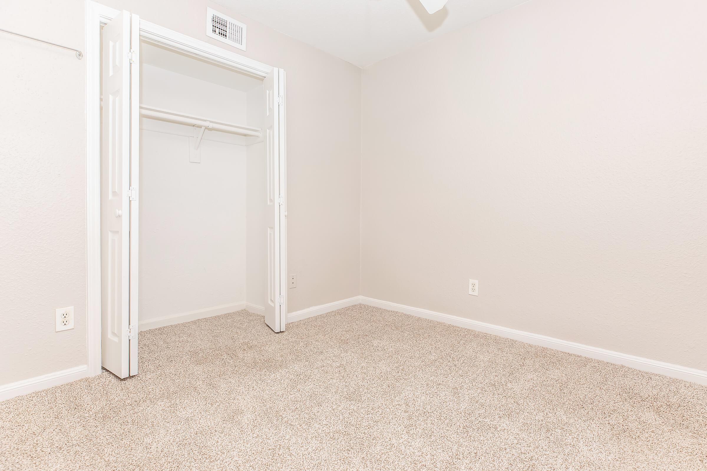 A vacant room with light beige walls and carpet. There is a closed white closet with double doors on the left side. The space appears clean and minimalistic, suitable for various uses. A ceiling fan is visible, and the room is well-lit.