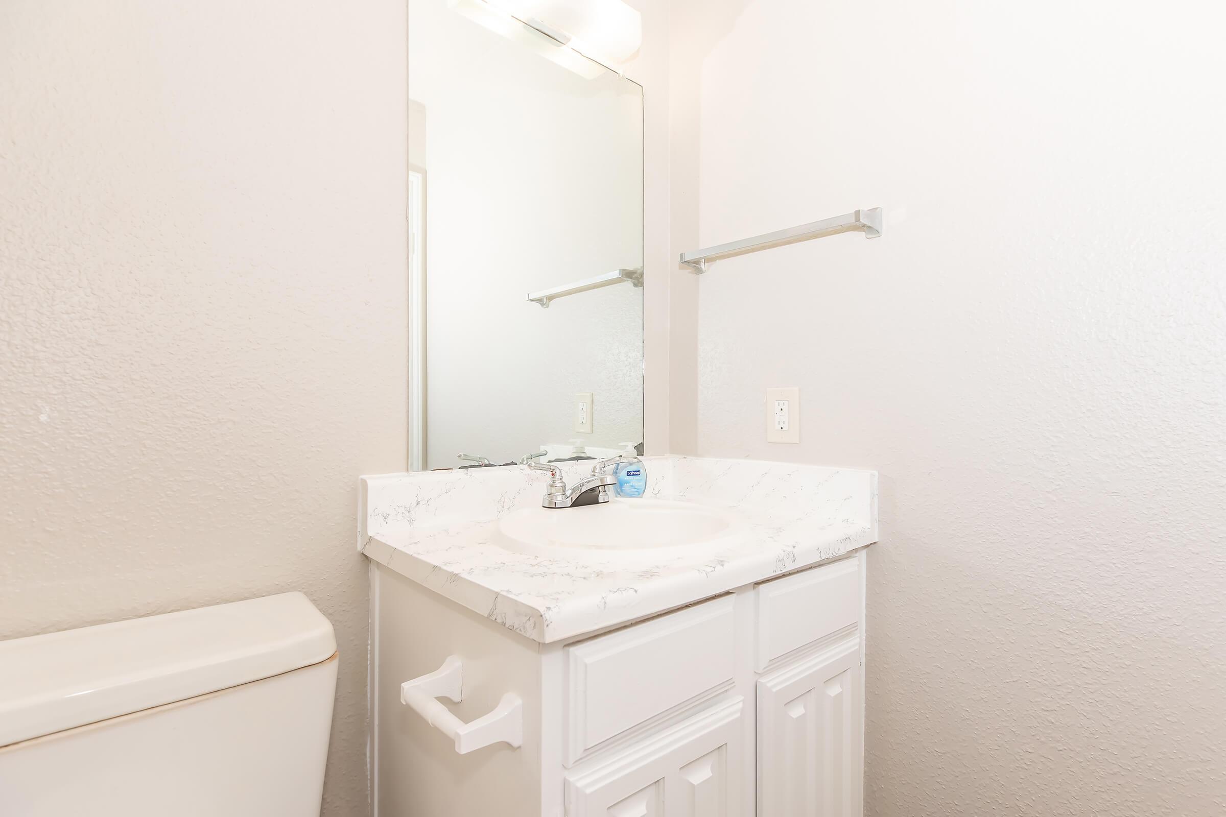 A clean, modern bathroom featuring a white marble countertop with a sink, a mirror above, and a towel rack. There is a white toilet on the left side against the light-colored wall, creating a bright and uncluttered space. Natural light illuminates the room, enhancing its fresh and airy feel.