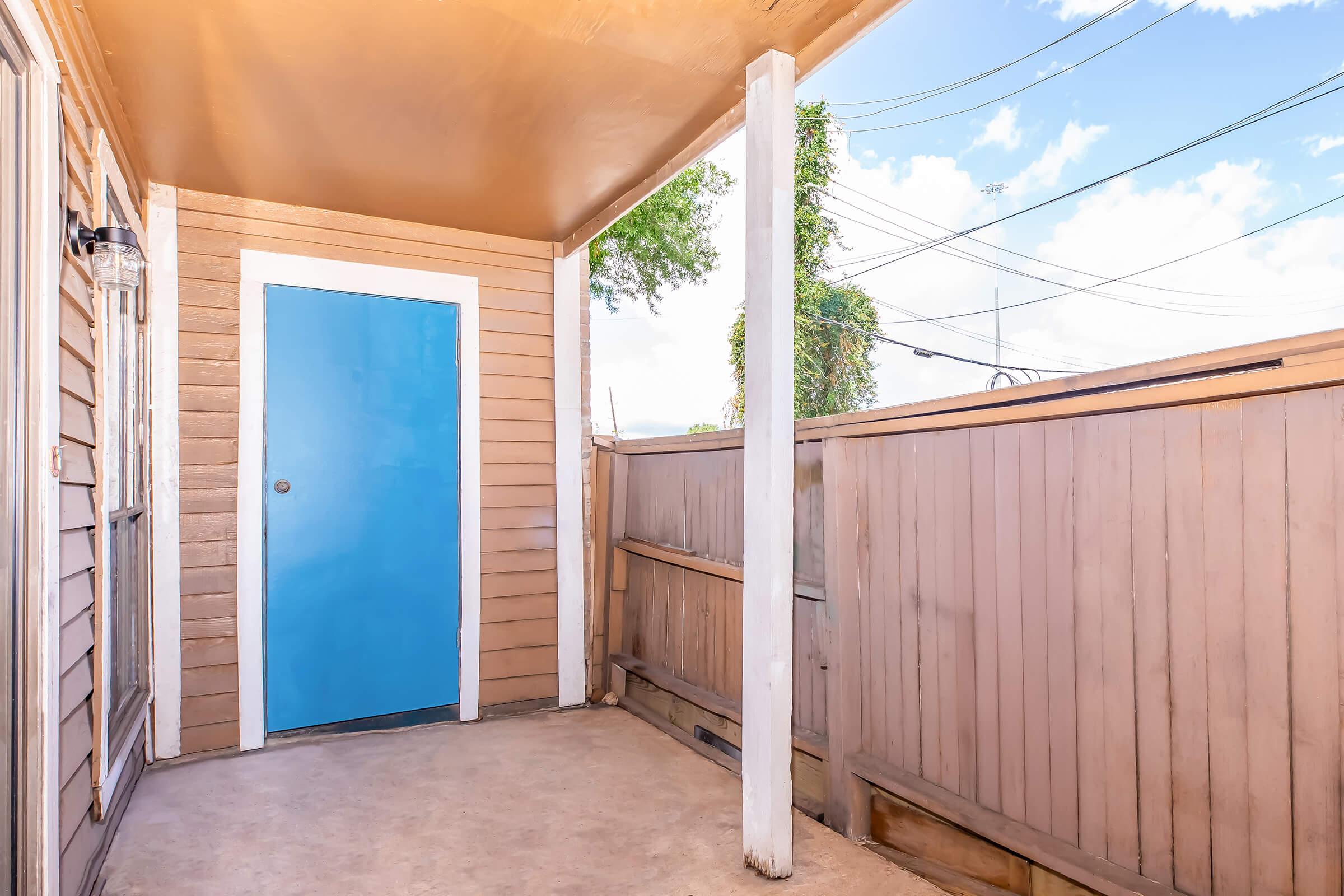 An empty porch area with a light-colored floor and walls. A bright blue door is centered at the back, with vertical wooden panels on either side and a roof overhead. The sky is visible above, with a few fluffy clouds and power lines in the background.