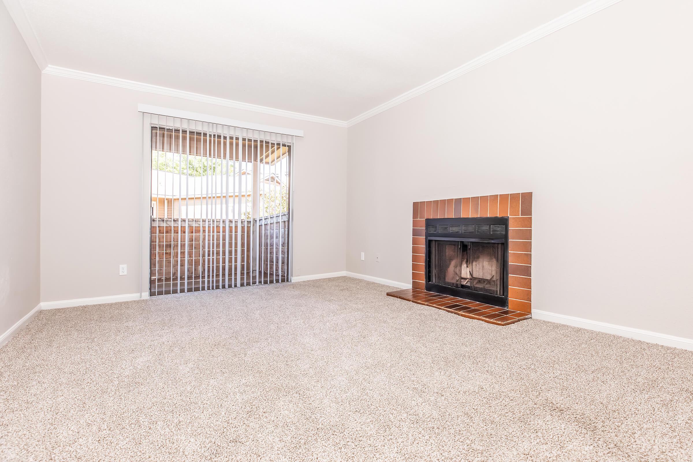 A spacious, clean living room featuring light-colored walls and a plush, beige carpet. A brick fireplace is positioned against one wall, and large sliding glass doors with vertical blinds provide natural light and open to an outdoor space. The room is empty and well-lit, creating a welcoming atmosphere.