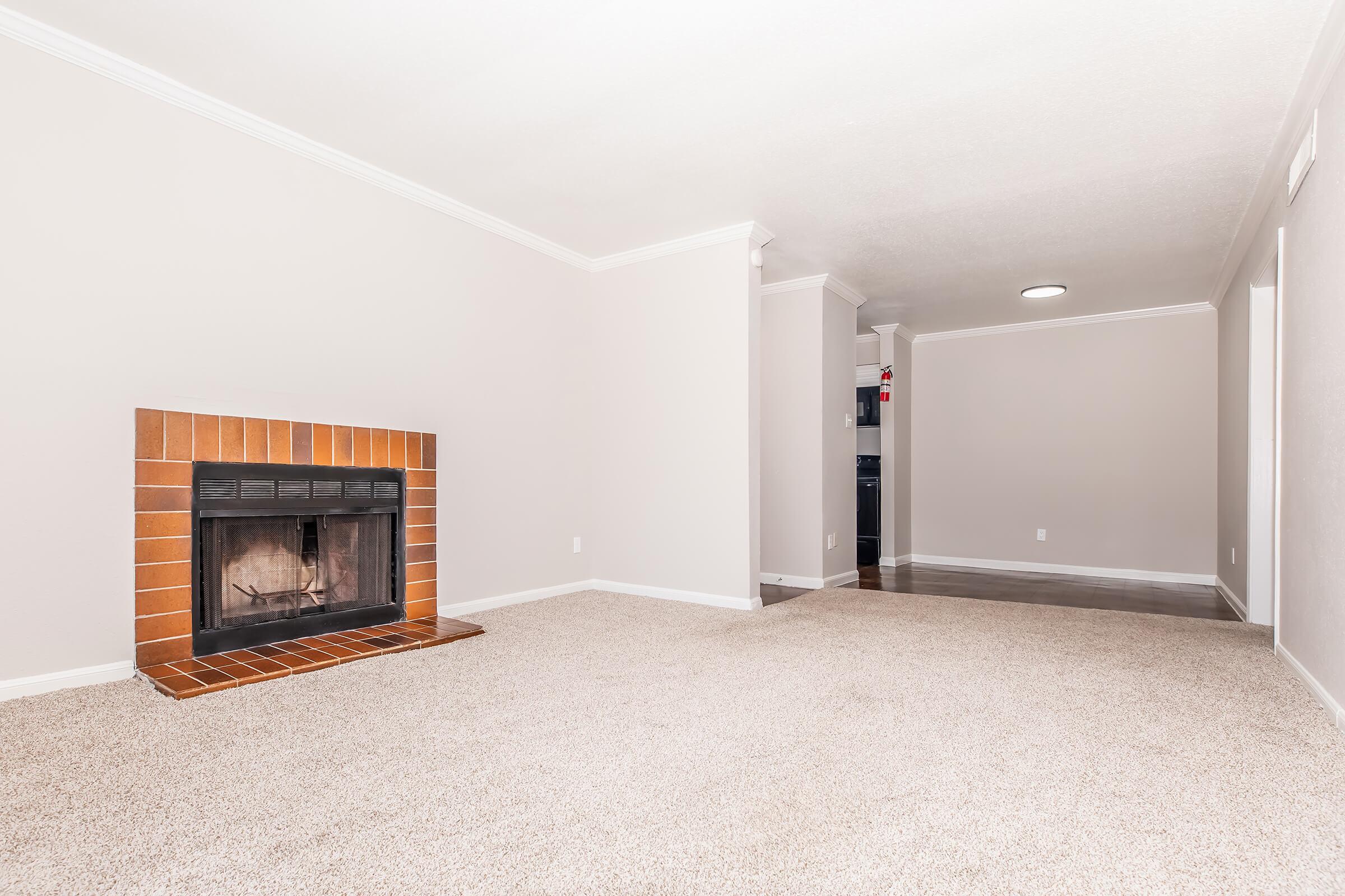 A spacious interior showing a living room with beige carpet and walls. There's a brick fireplace on the left with a dark metal grate. Adjacent to the fireplace is an open area leading to a kitchen, visible in the background on the right, with a modern design and overhead lighting.