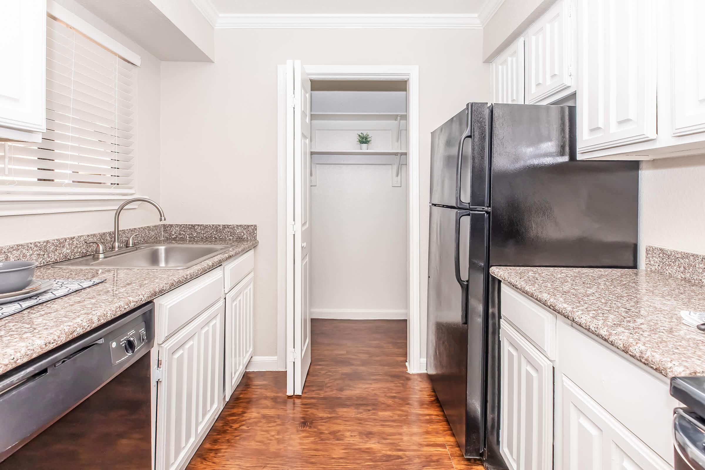 A modern kitchen featuring white cabinets, a granite countertop, and stainless steel appliances. On the left, a sink and dishwasher; on the right, a black refrigerator. A doorway leads to a small pantry with a plant on a shelf, and the flooring is a warm wood finish. Natural light comes from a window above the sink.