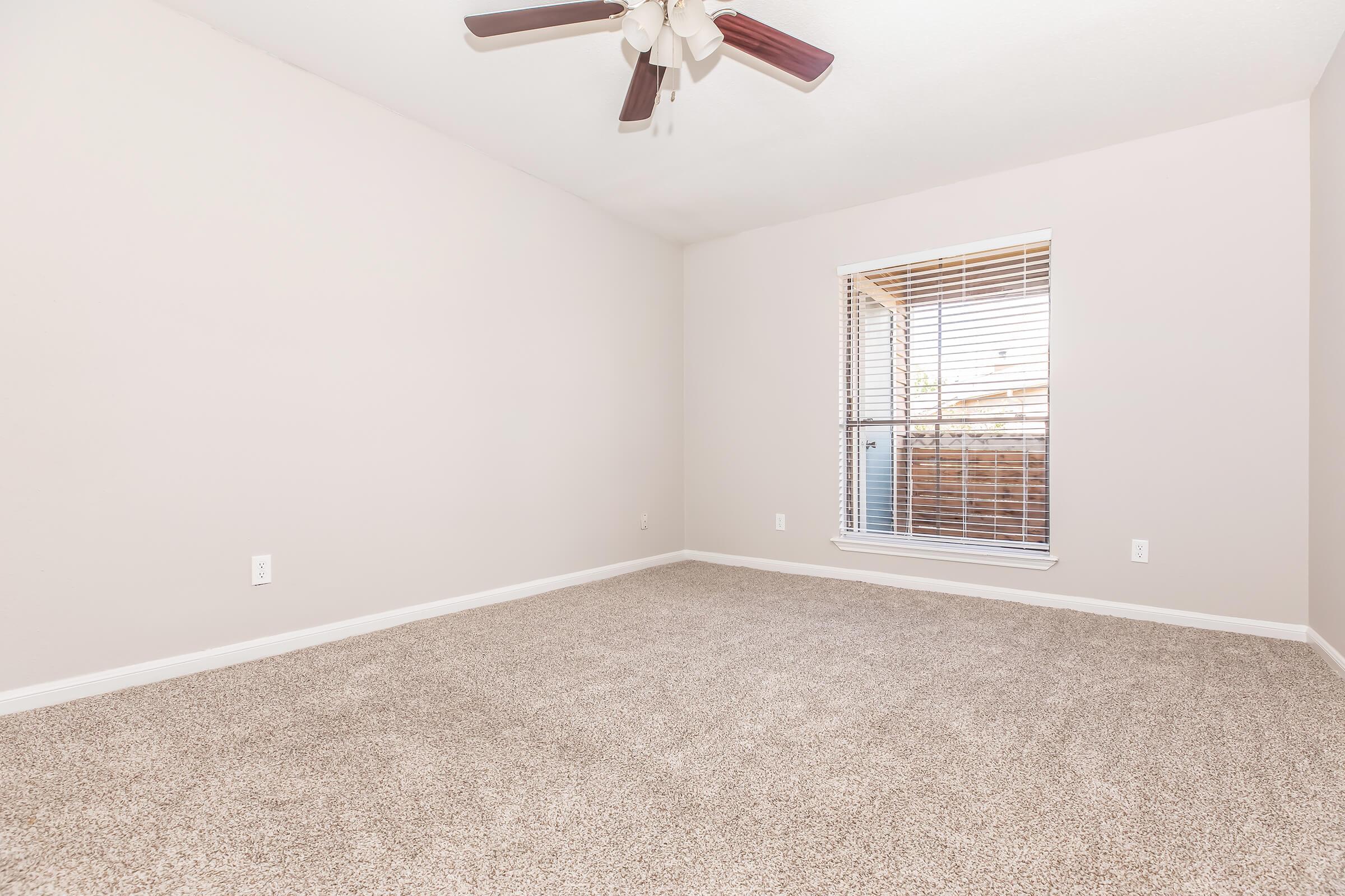 A spacious, empty room with light-colored walls and a ceiling fan. The floor is covered in beige carpet, and there is a window with blinds that lets in natural light. The overall ambiance is bright and inviting, perfect for various interior design possibilities.