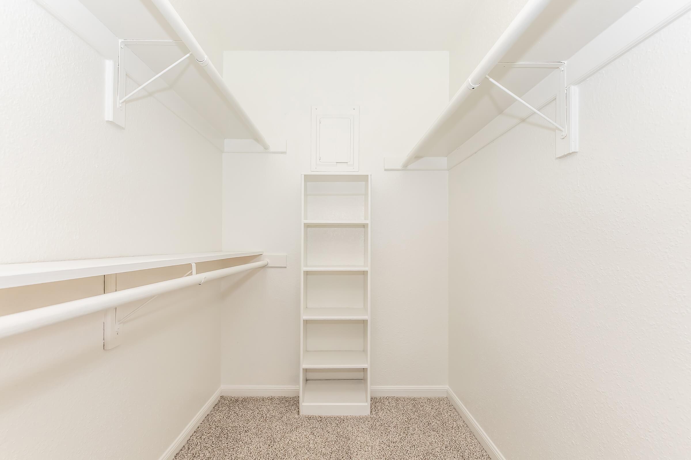 A spacious, empty walk-in closet featuring white walls and shelving. There are two hanging rods on either side and a central white shelving unit, all set against a carpeted floor. The overall design is minimalist and clean, providing plenty of storage potential.