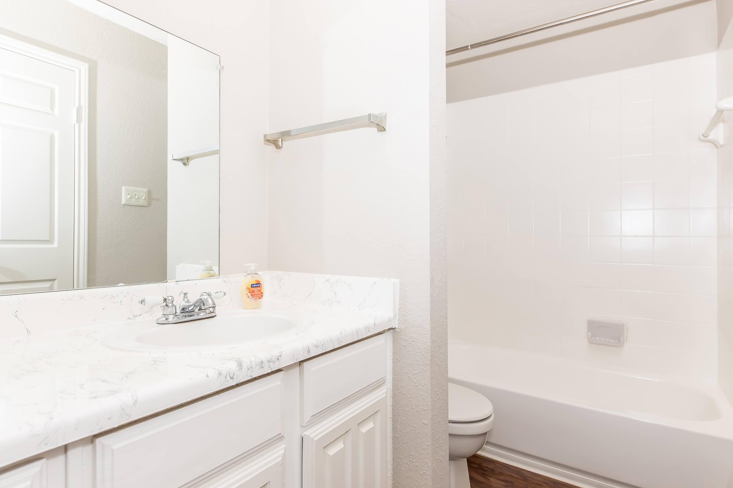 A bright bathroom featuring a white countertop with a sink and silver faucet, a wall-mounted towel bar, and a clean bathtub with a shower. The walls are painted in a light color, and a mirror reflects the space. Natural light enhances the tidy appearance.