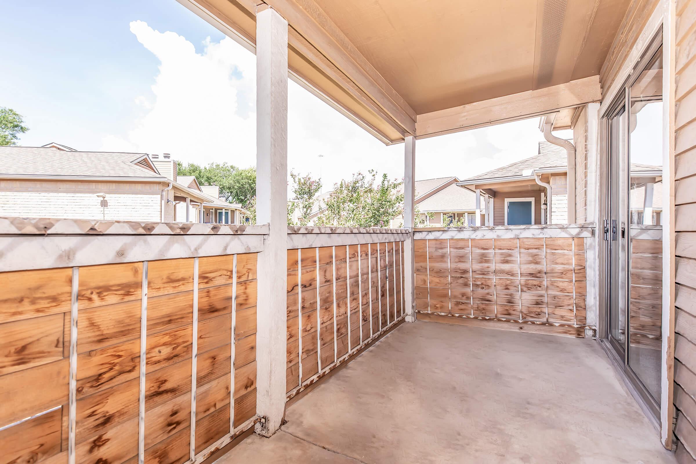 A covered balcony with a concrete floor and wooden railing, featuring a view of nearby houses and blue sky with clouds. The space is well-lit and inviting, ideal for relaxation or enjoying the outdoors.