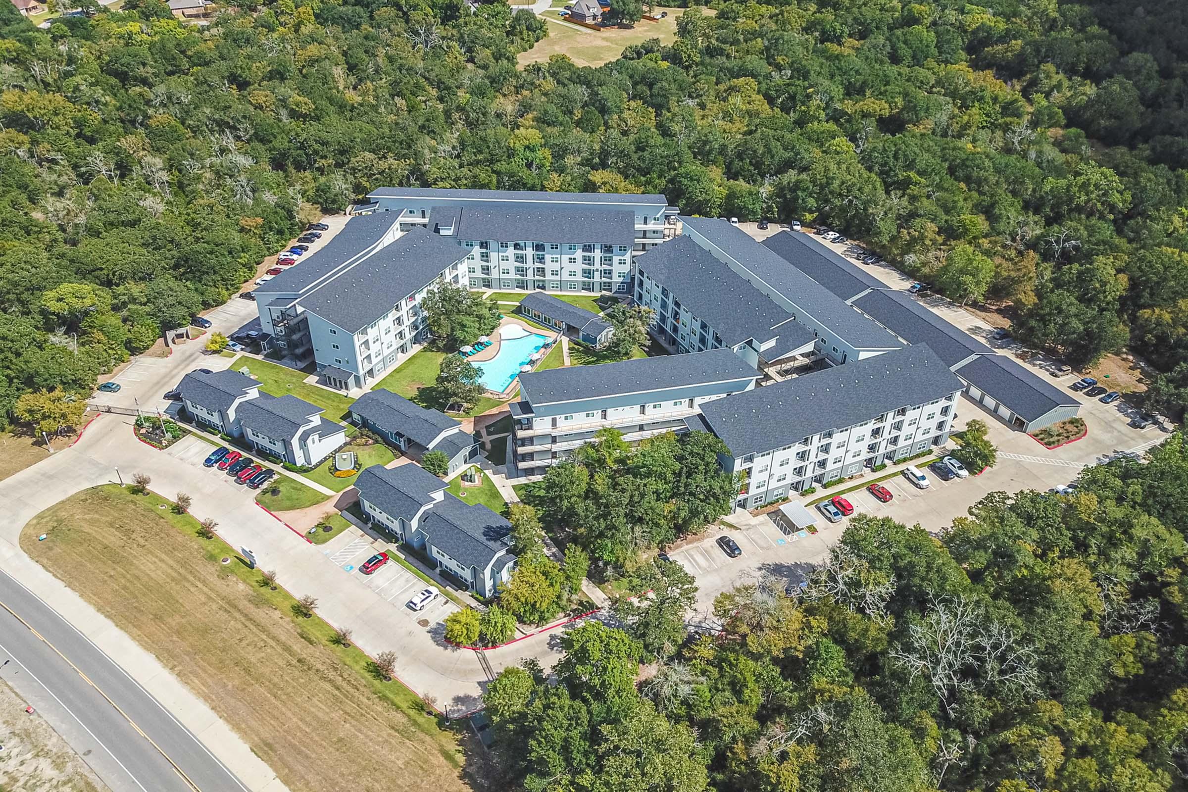 Aerial view of a hotel complex surrounded by trees, featuring multiple building structures in a U-shape, a central pool area, and parking spaces. The landscape includes grassy areas and pathways, with a road leading to the entrance. The setting appears peaceful and well-maintained.