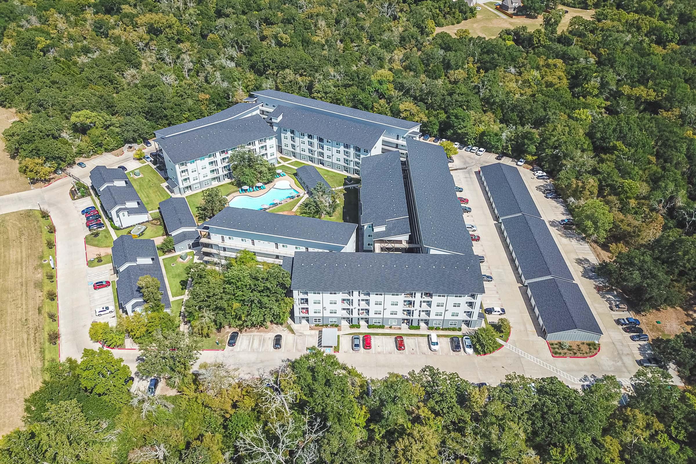 Aerial view of a hotel complex surrounded by trees, featuring multiple buildings arranged around a central pool area. Parking spaces are visible in front of the buildings, and lush greenery surrounds the property. The setting appears tranquil and inviting, typical of a resort-style accommodation.