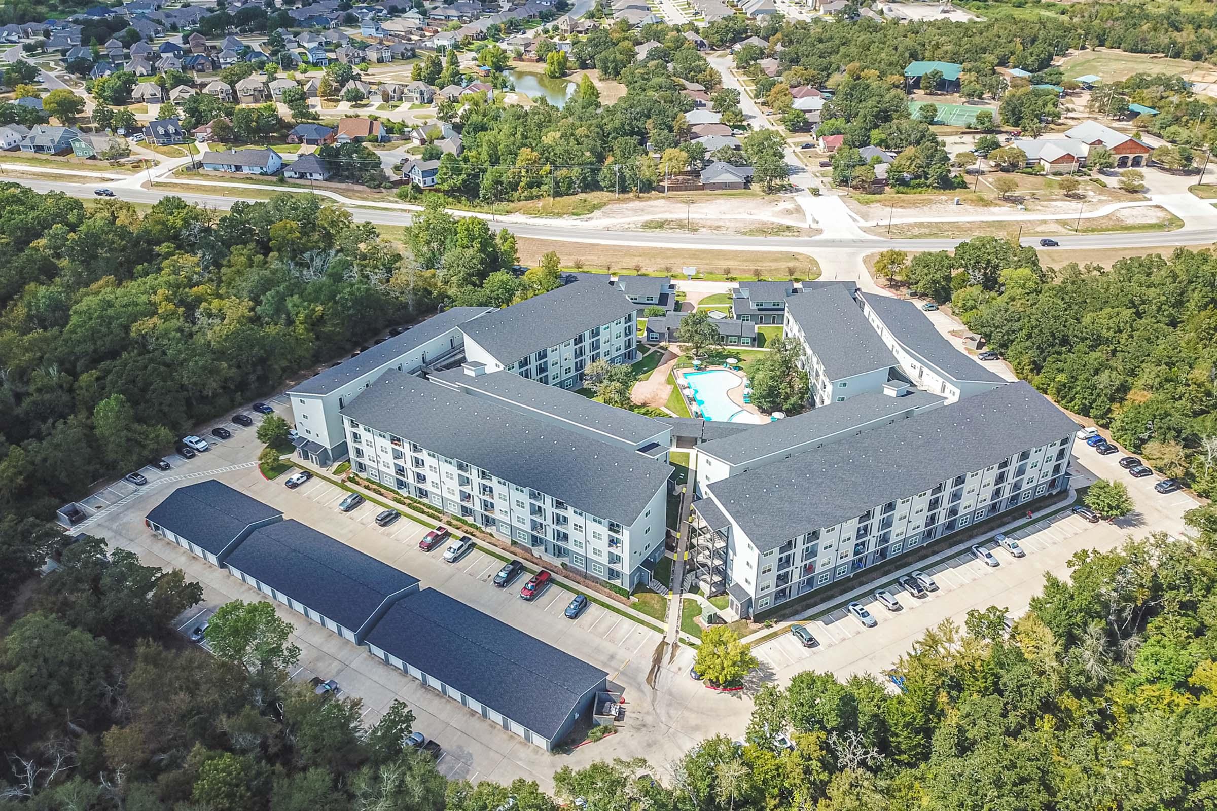 Aerial view of a multi-story apartment complex with a swimming pool and parking lot, surrounded by trees and residential areas. The layout features several buildings in a U-shape, with additional covered parking spaces visible in the foreground, and roads leading to nearby neighborhoods.