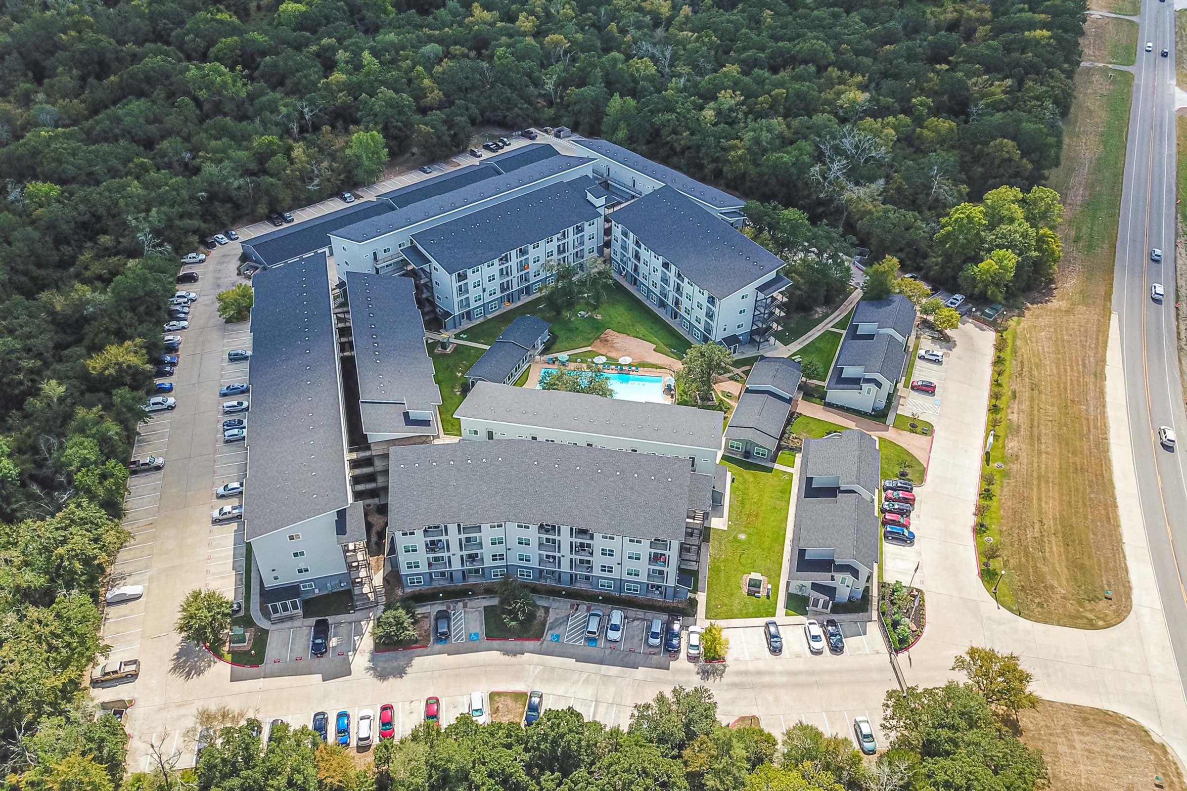 Aerial view of a multi-story apartment complex with a central pool area surrounded by green landscaping. Several parking lots are visible, with numerous parked cars. The surrounding area is filled with trees, and a road runs alongside the property.