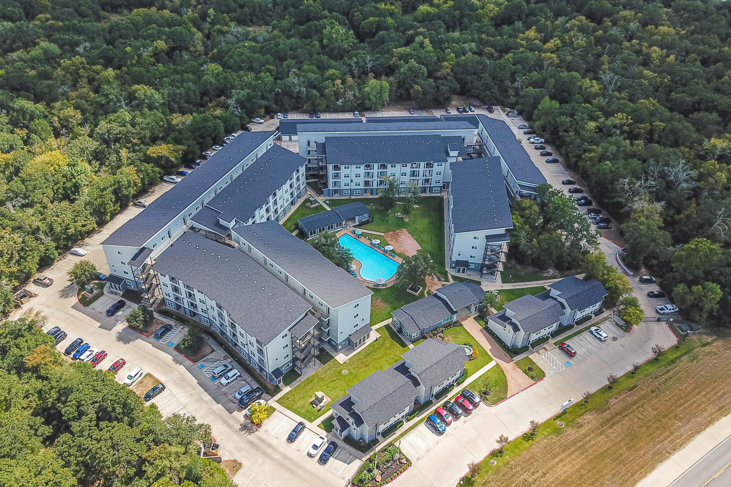 Aerial view of a residential complex surrounded by trees, featuring multiple buildings arranged around a central pool area. The complex includes green lawns, parking spaces, and pathways, with a nearby road lined with trees.