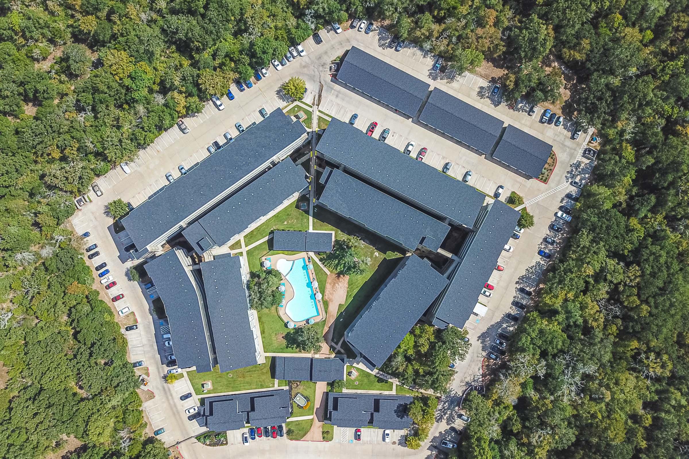 Aerial view of a complex featuring multiple buildings arranged in a U-shape around a central swimming pool. Surrounding the buildings are green trees and a parking lot filled with vehicles. The scene showcases a blend of nature and residential architecture in a tranquil setting.