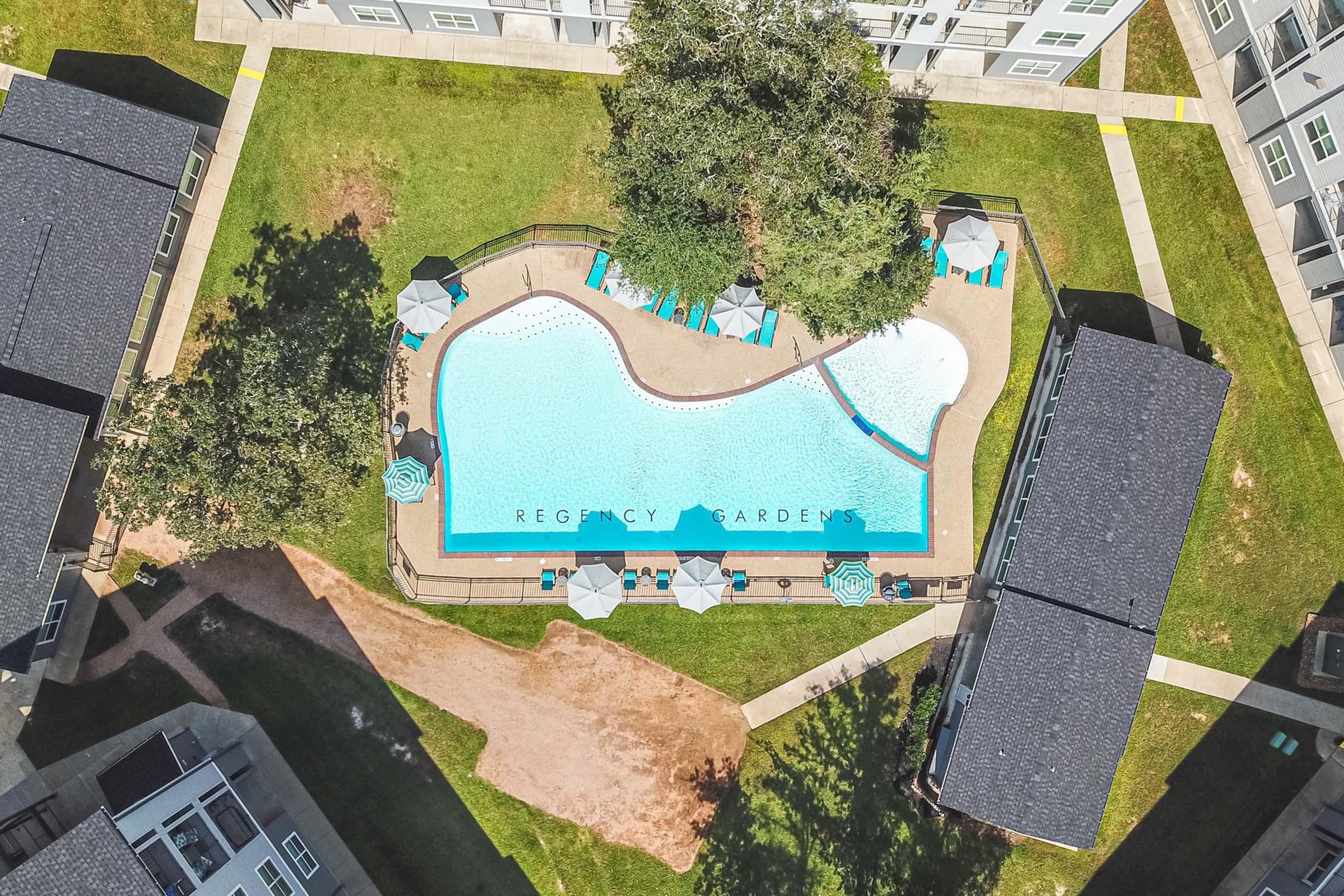 Aerial view of a residential complex featuring a large, irregularly shaped swimming pool surrounded by green grass. Lounge chairs with blue-and-white umbrellas are set up around the pool, and two low buildings are visible adjacent to the pool area. The text "REGENCY GARDENS" is superimposed over the pool.