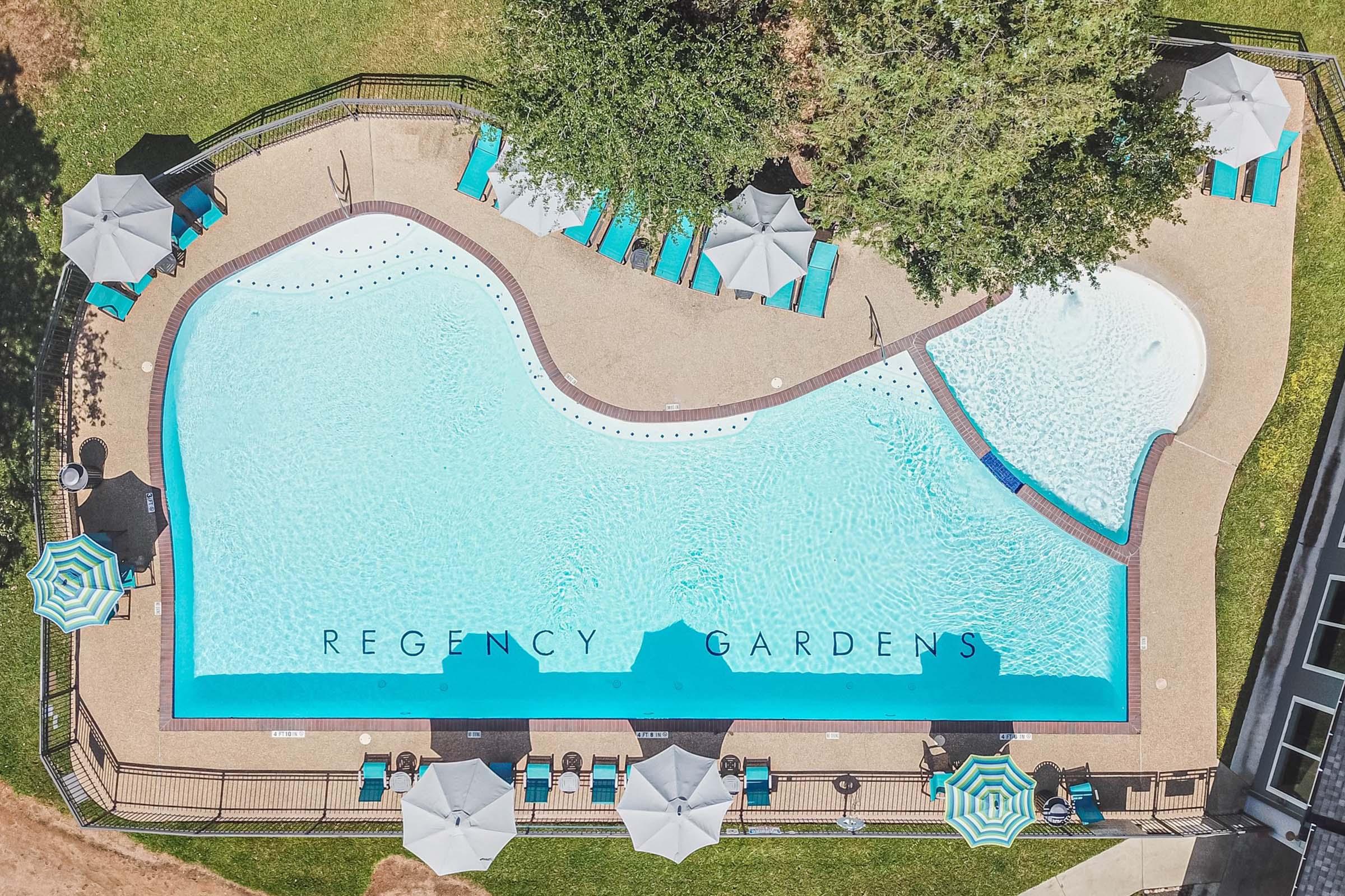 Aerial view of a swimming pool at Regency Gardens, featuring blue water, lounge chairs, and umbrellas. The pool is surrounded by greenery, with distinct areas for relaxation and recreation.