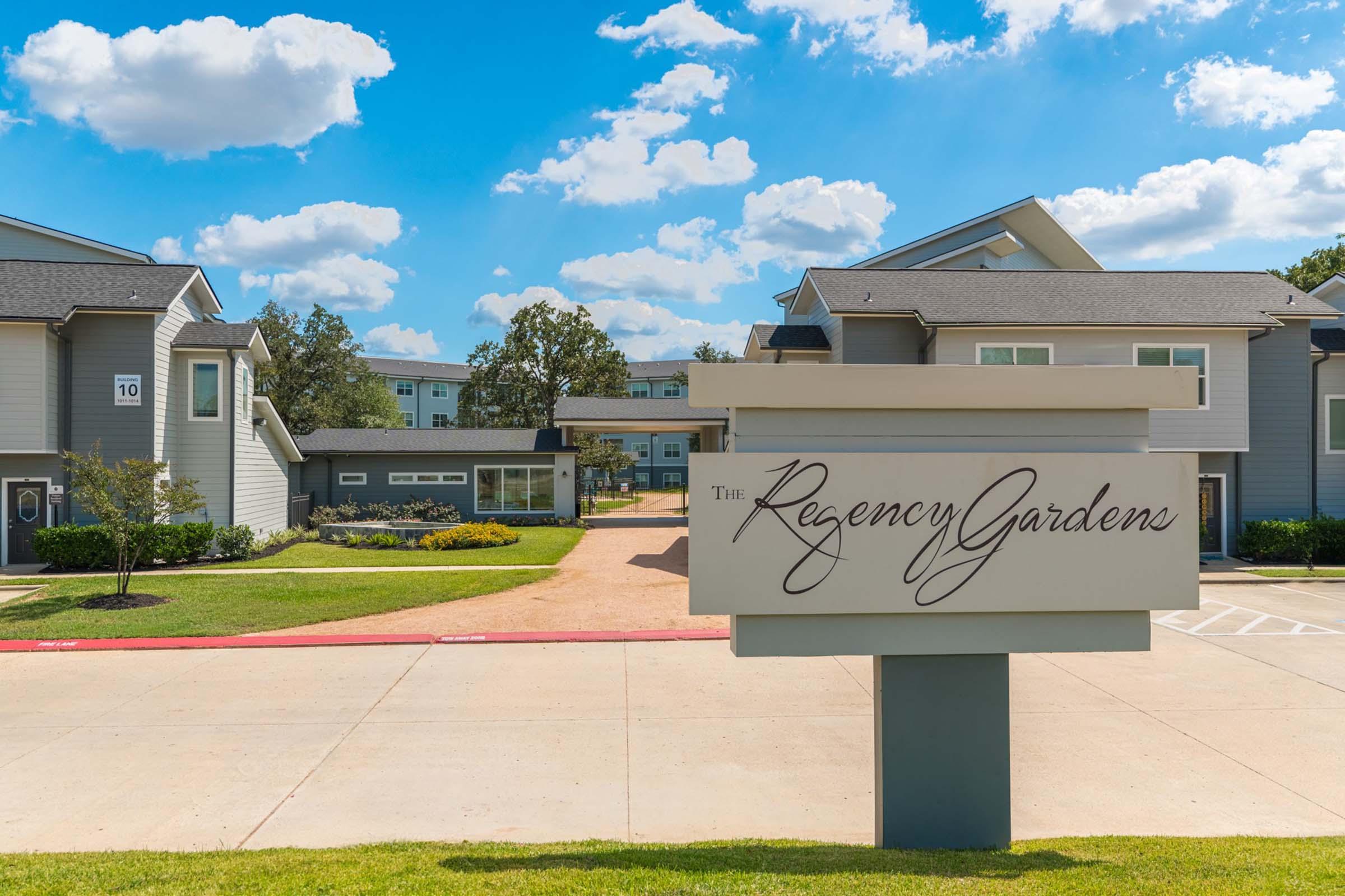 Sign for "The Regency Gardens" in front of a residential complex. The building features modern architecture with gray siding and landscaped greenery. Clear blue sky with fluffy white clouds overhead, creating a bright and inviting atmosphere.