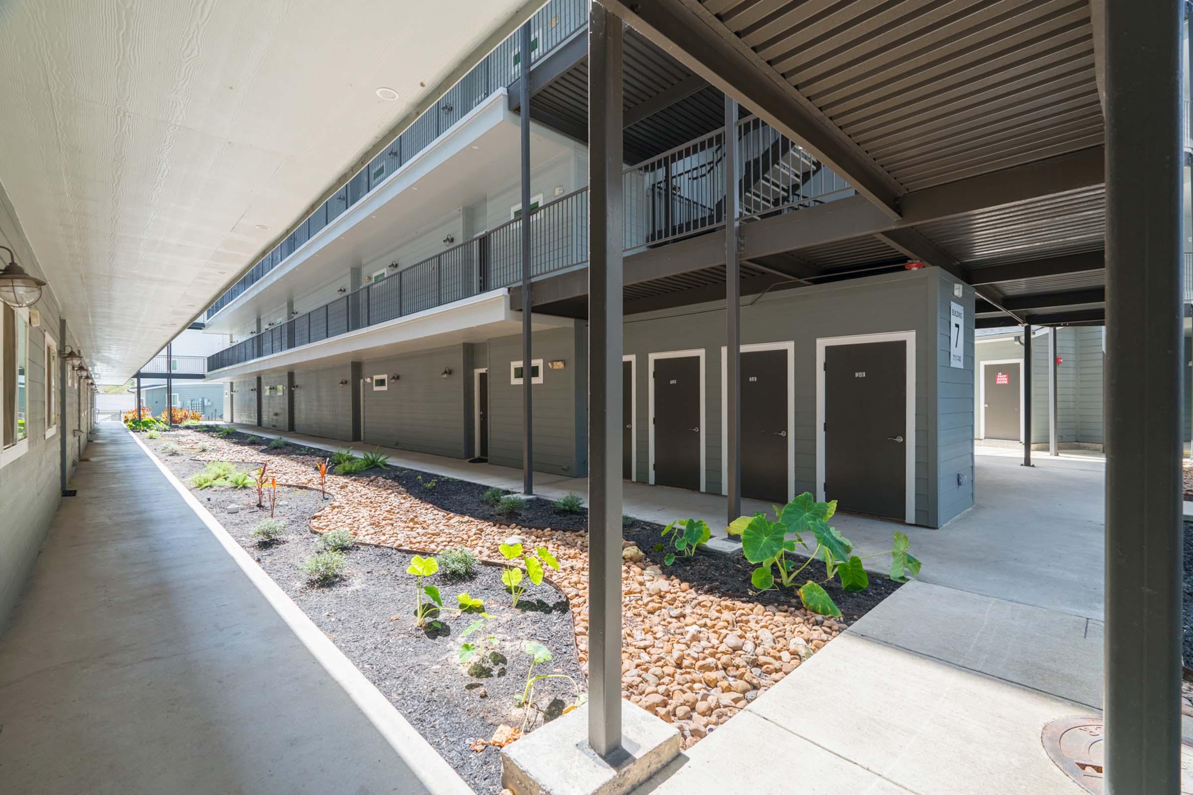 A modern multi-story building with a courtyard. The image shows a walkway lined with plants and stones, leading to rows of dark doors. The structure features multiple floors with balconies and a clean, contemporary design. Sunlight illuminates the scene, creating a bright and welcoming atmosphere.