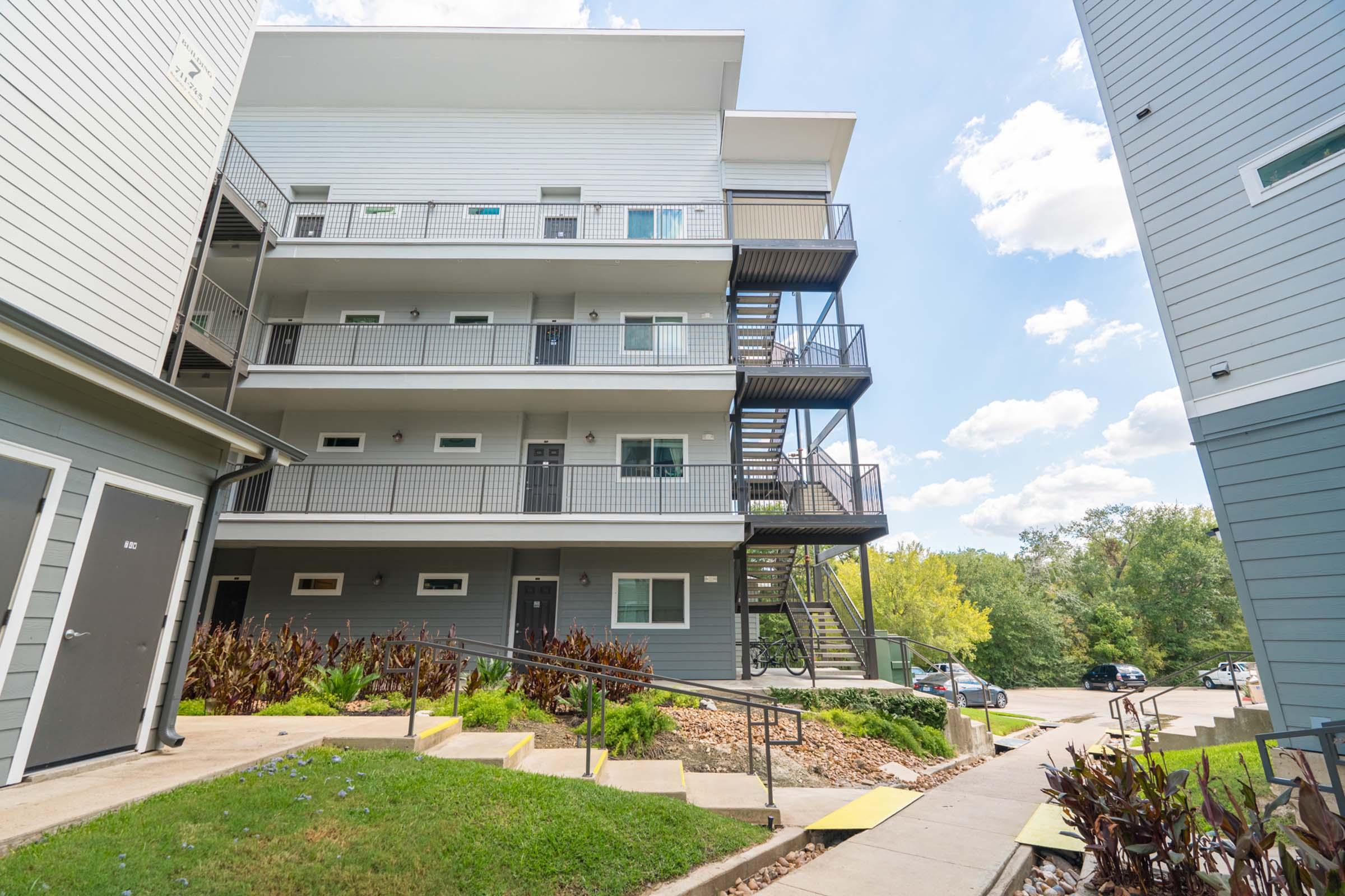 A multi-story residential building with a modern design. The exterior features light gray siding and black metal staircases. Surrounding the building are well-maintained pathways and landscaping. A parking area is visible in the background, along with trees and a clear blue sky.