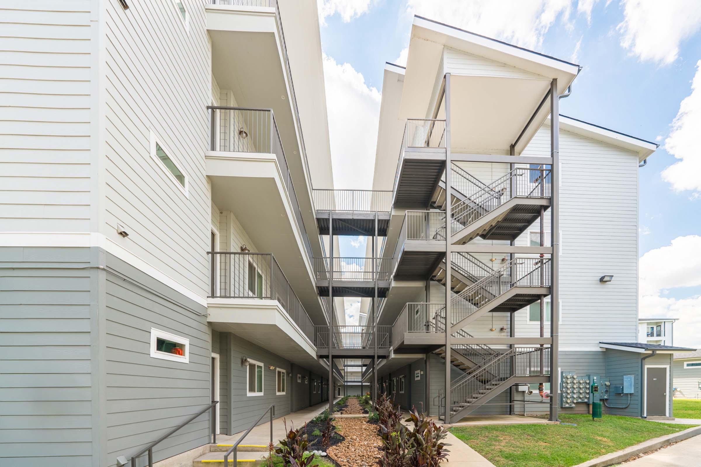 View of an apartment complex showcasing modern architecture. The image features multiple levels with balconies, metal staircases, and landscaped areas between the buildings. The sky is partly cloudy, adding to the bright, open atmosphere of the setting.