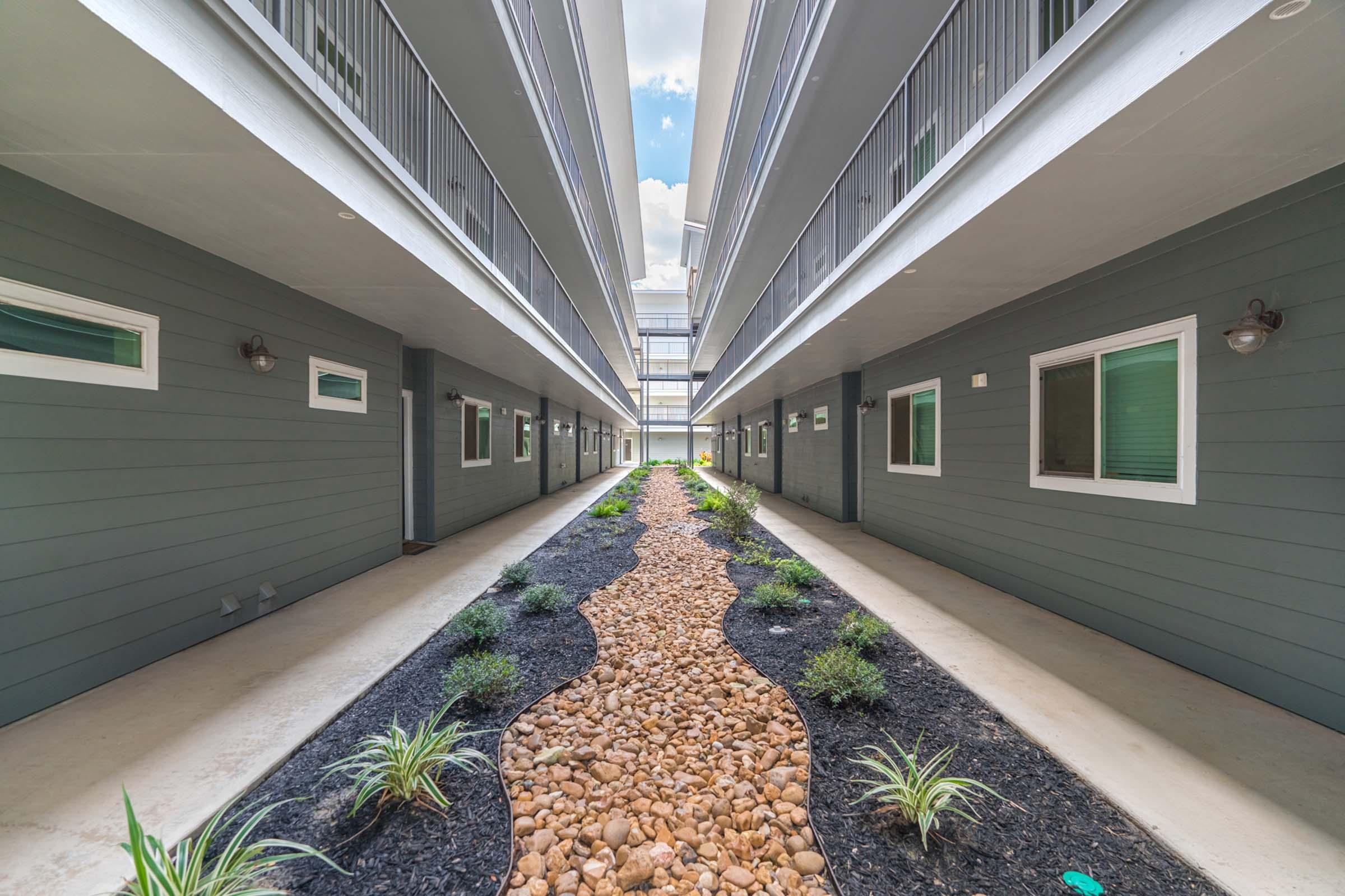 A view of a modern apartment complex interior, showcasing a stone pathway lined with small plants and greenery. The buildings feature gray exteriors with multiple windows and balconies, creating a symmetrical layout along the corridor. Bright blue sky visible above.