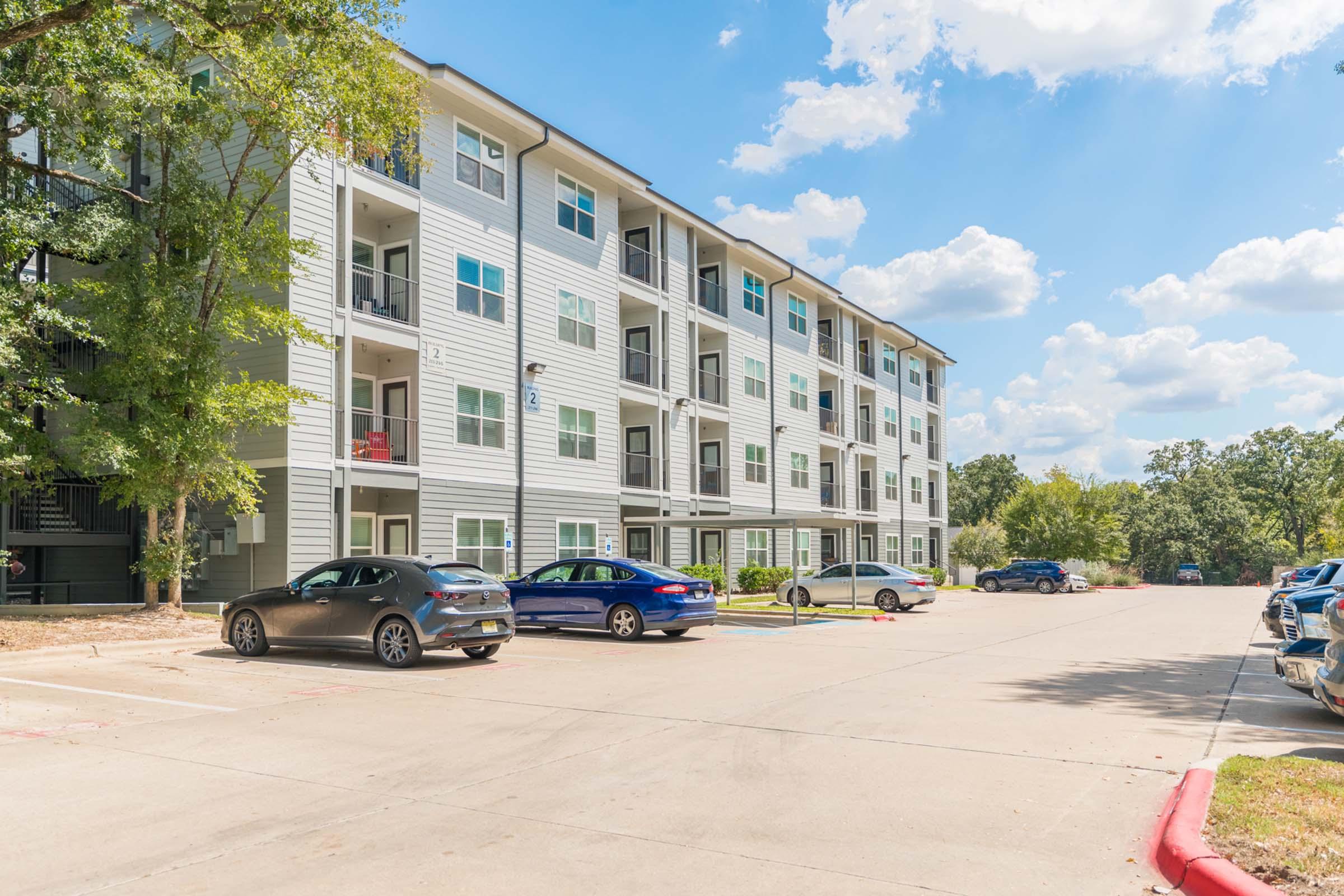 A modern multi-story apartment building with balconies, surrounded by a parking lot filled with cars. The scene is set on a sunny day with a clear blue sky and scattered clouds. Trees and greenery are visible in the background, contributing to a pleasant residential atmosphere.