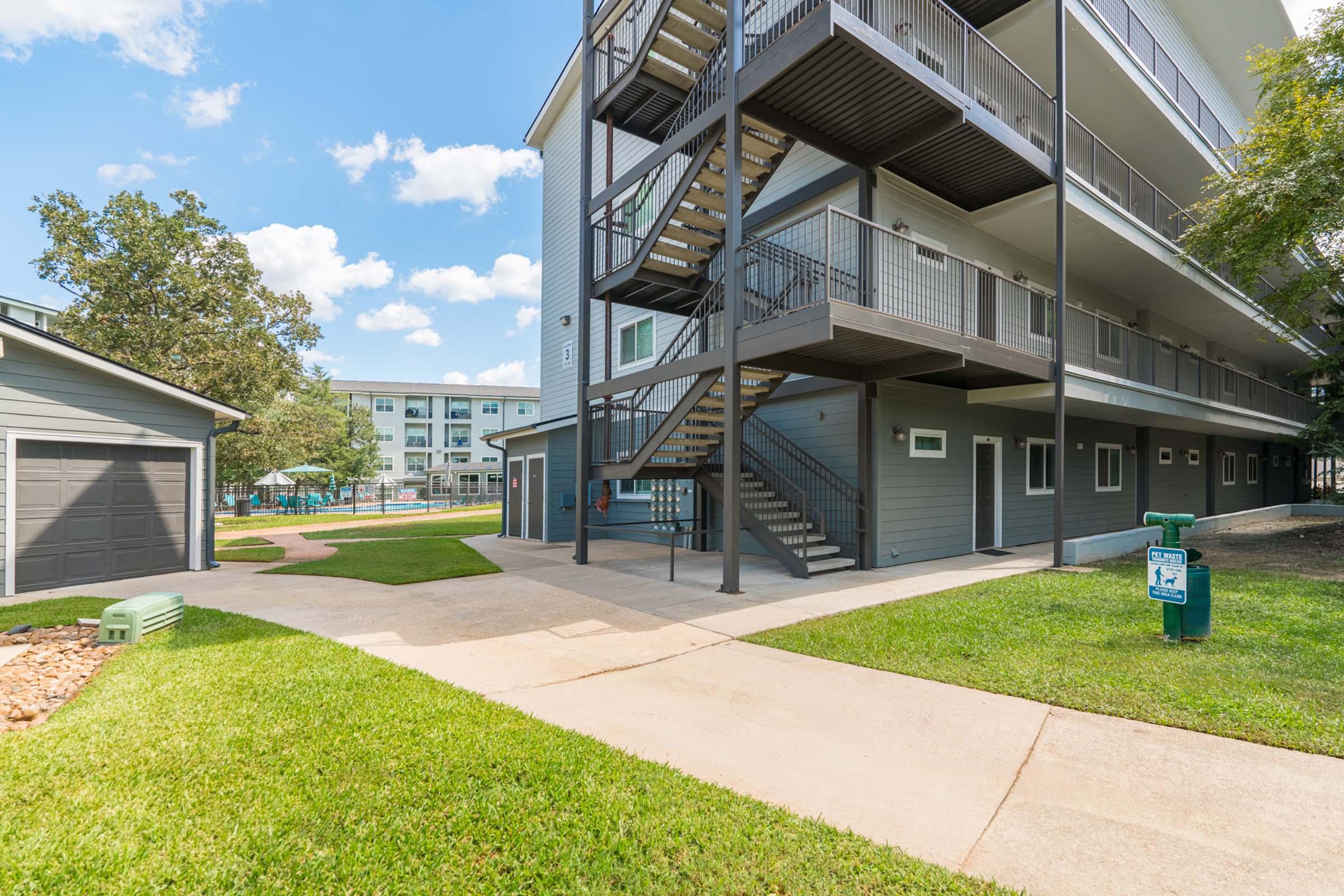 A modern multi-story building with metal outdoor stairs, grass pathways, and landscaped areas. In the background, a pool area is visible along with other residential buildings. The setting features clear blue skies and sunlight illuminating the space.