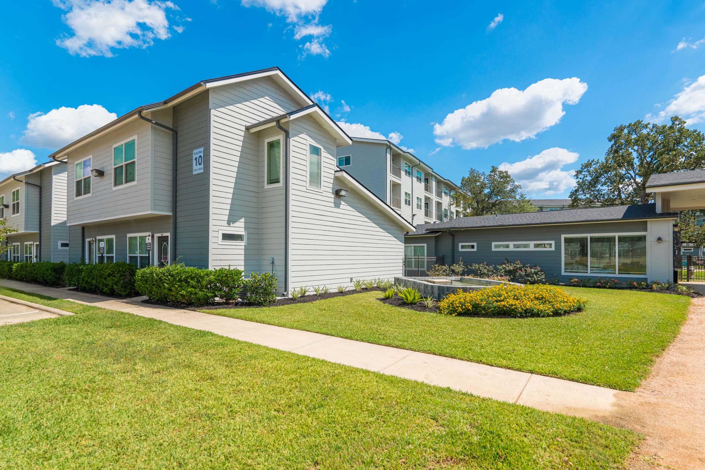 Two modern apartment buildings are situated in a well-maintained landscape with green grass and colorful flower beds. The sky is bright blue with fluffy white clouds. A paved sidewalk runs alongside the buildings, enhancing the inviting and pleasant atmosphere of the residential complex.