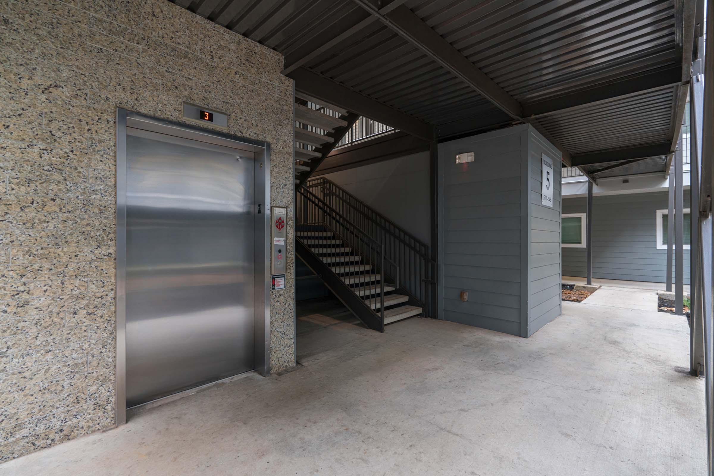 A modern apartment building entrance featuring an elevator on the left, a stairway leading up, and a wall with the number 5. The space is well-lit with concrete flooring and a mix of stone and siding materials.