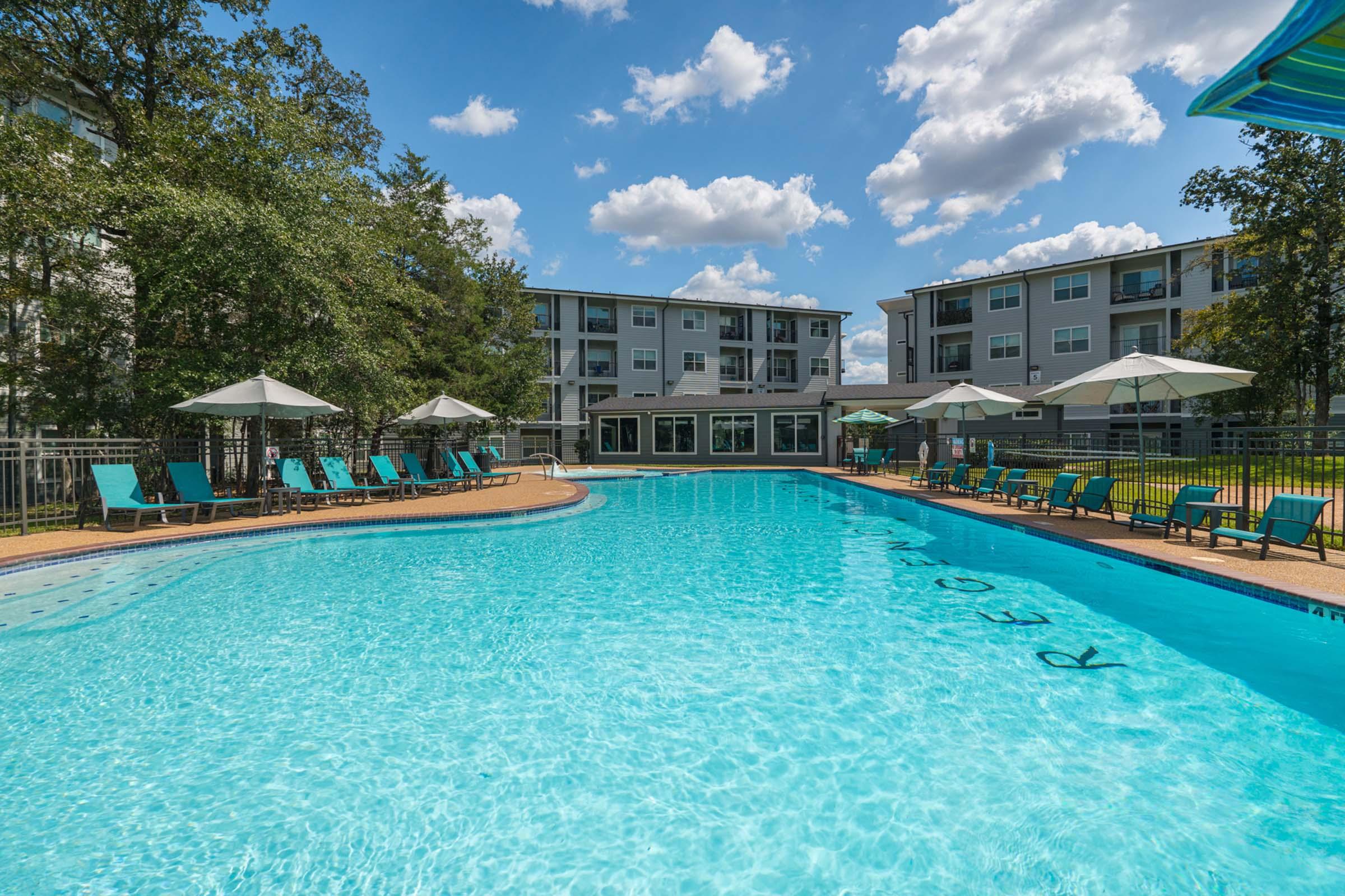 A bright, sunny outdoor swimming pool surrounded by lounge chairs and umbrellas. In the background, there are two multi-story apartment buildings against a backdrop of blue sky and fluffy clouds. The pool area is well-maintained and inviting, perfect for relaxation and leisure activities.