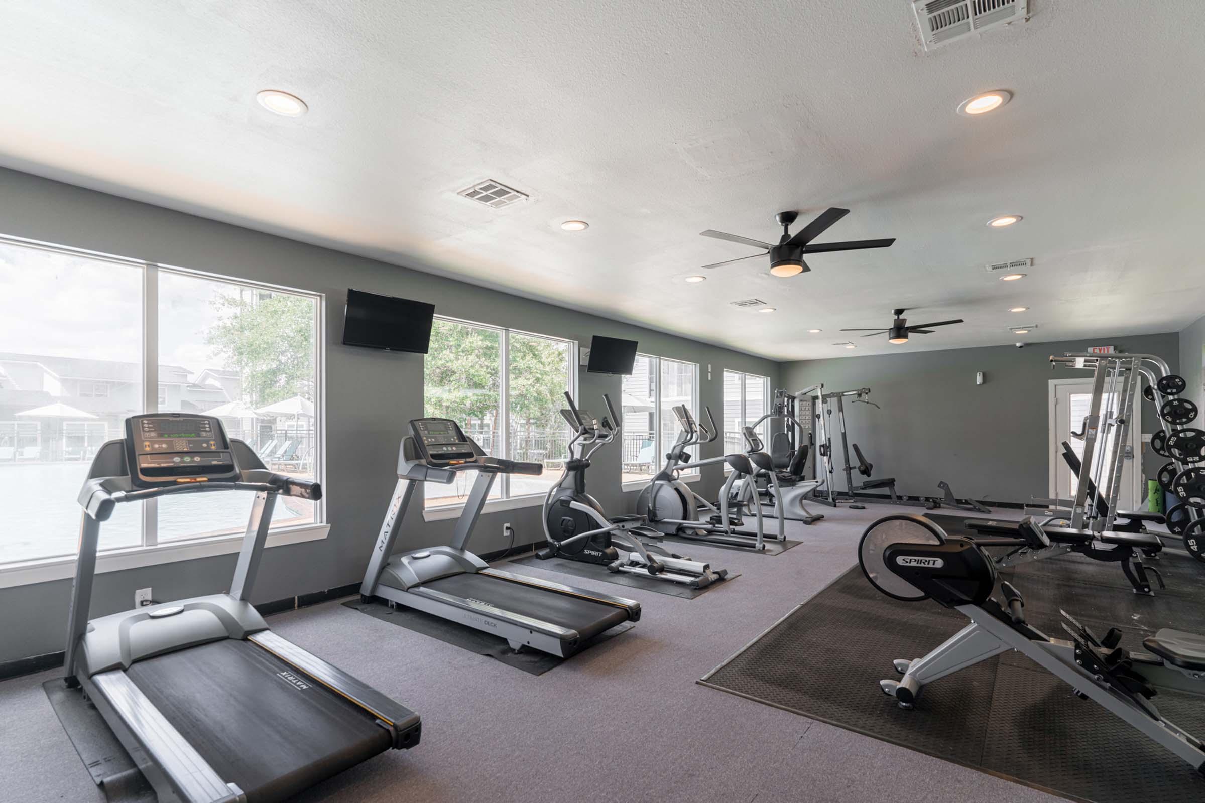 A modern gym interior featuring multiple treadmills, ellipticals, and weight training equipment. Large windows allow natural light to fill the space, and ceiling fans enhance comfort. Two wall-mounted televisions are visible. The flooring is carpeted, creating a welcoming exercise environment.