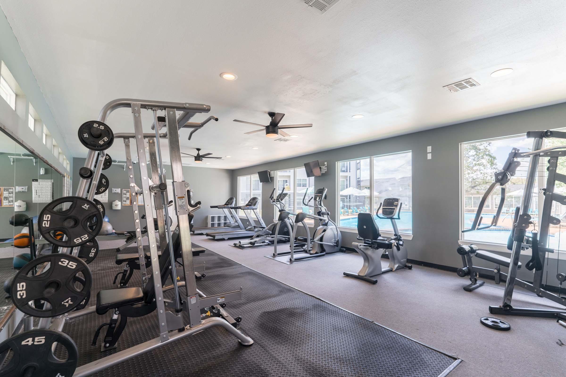 A modern gym featuring various exercise equipment including free weights, a weightlifting machine, and cardio machines. The gym is well-lit with large windows showing an outdoor area, and has a clean, organized layout with a rubber flooring surface.